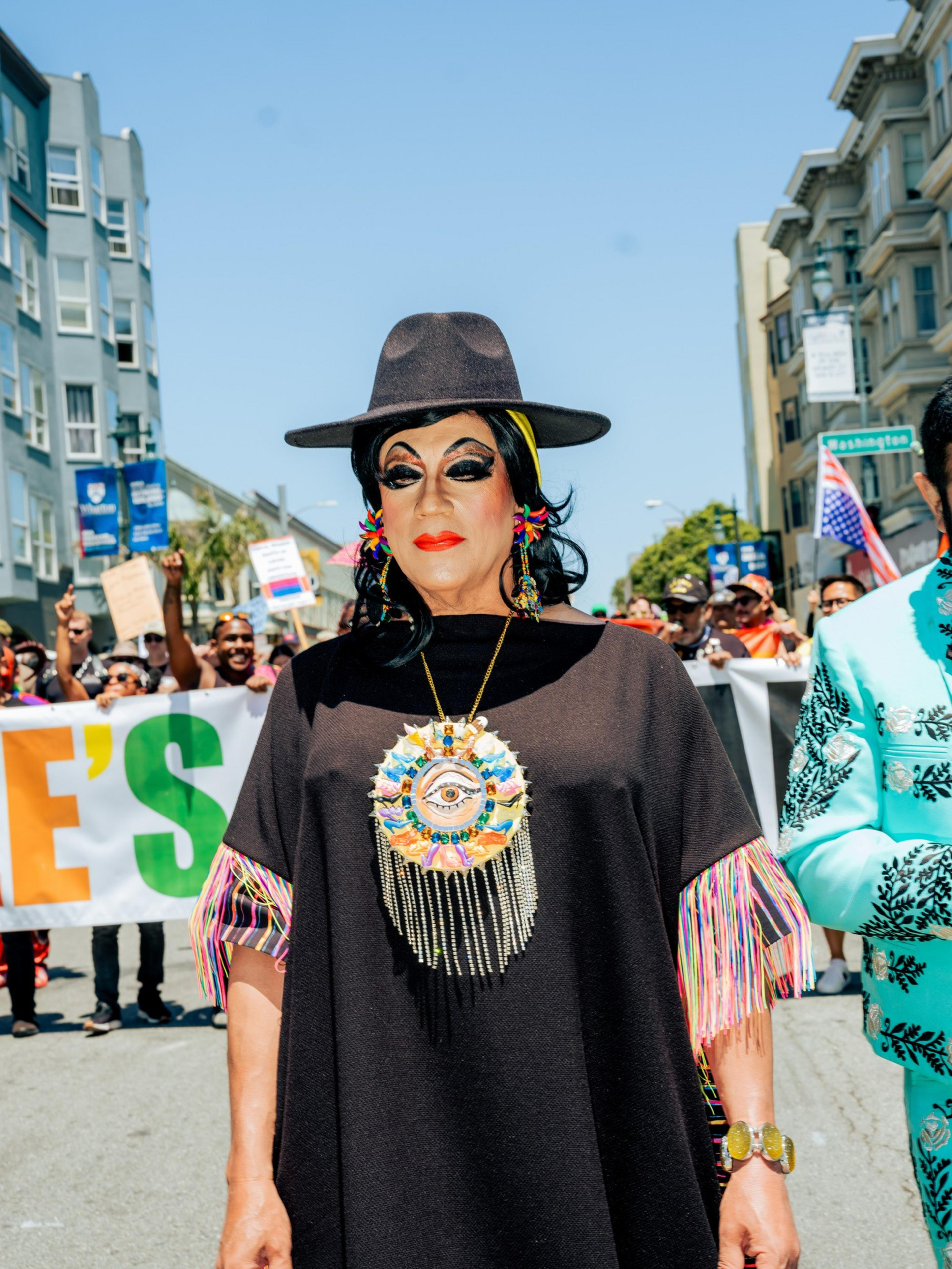 A person wearing vibrant makeup, a black wide-brimmed hat, and a necklace with an eye design, stands in front of a colorful parade under a clear sky.