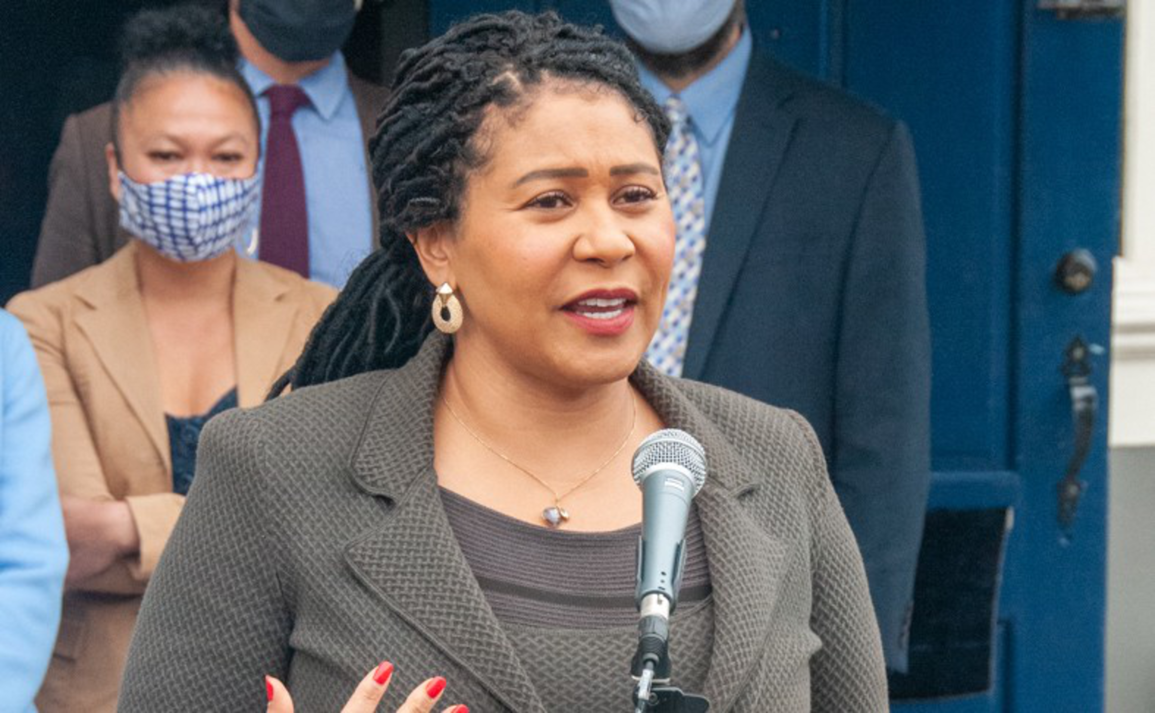 A woman with braided hair speaks into a microphone, wearing a grey textured blazer, with three masked people standing behind her.