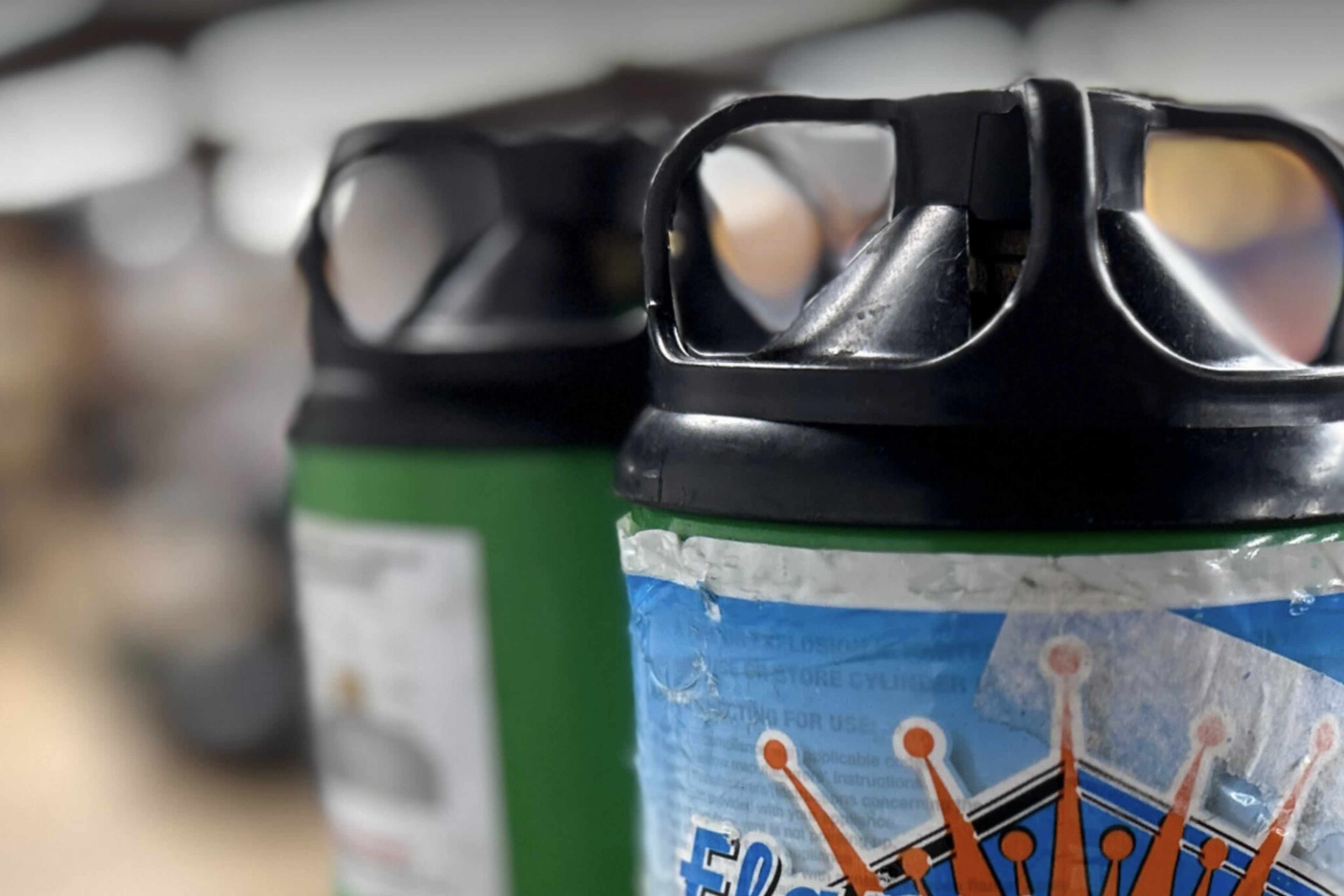 The image shows two green Flame King refillable propane cylinders with black caps, standing close together on a wooden surface in a blurred indoor setting.