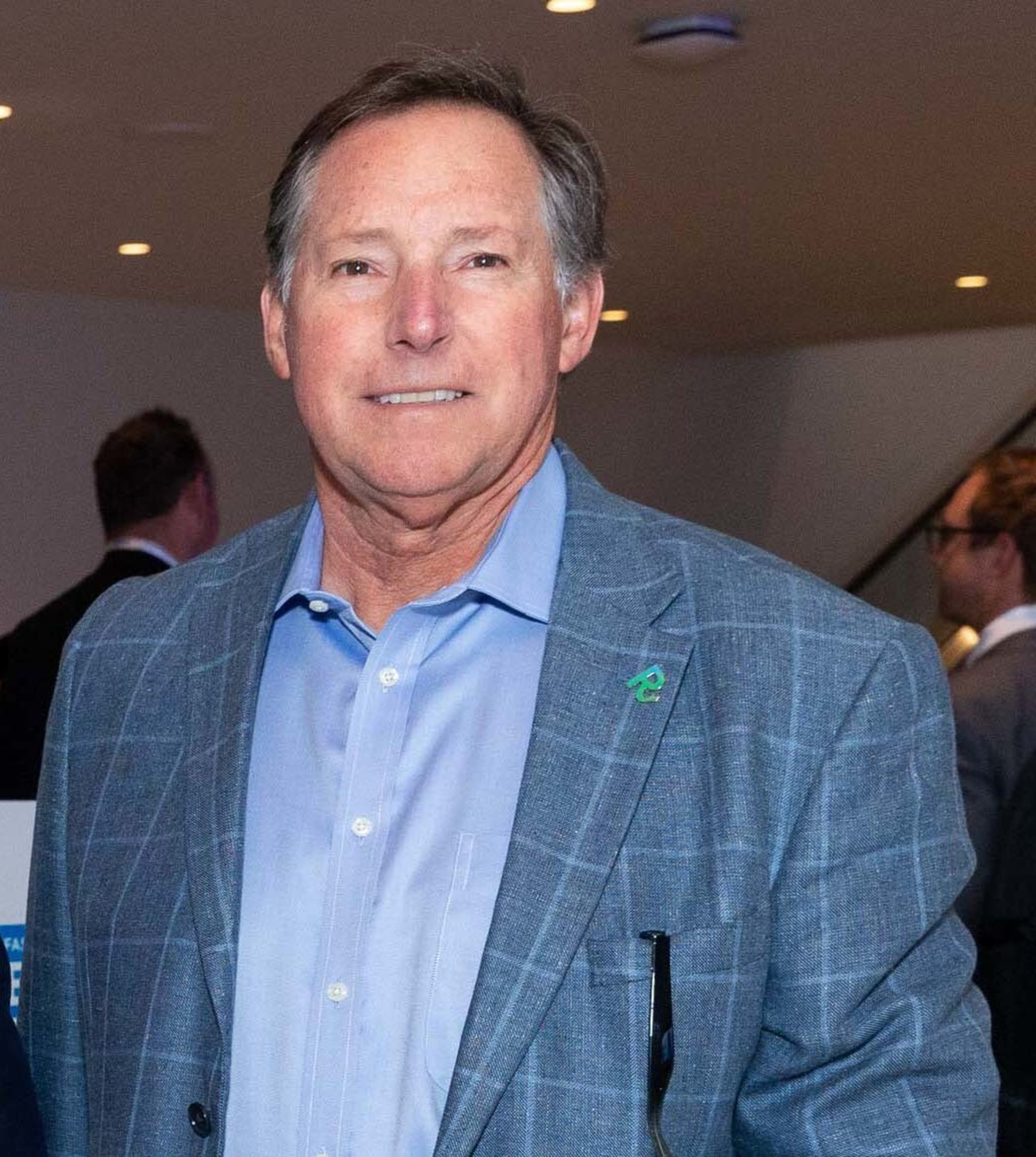 A middle-aged man wears a light blue button-up shirt and a blue plaid blazer with a small green pin on the lapel, standing indoors with people blurred behind him.