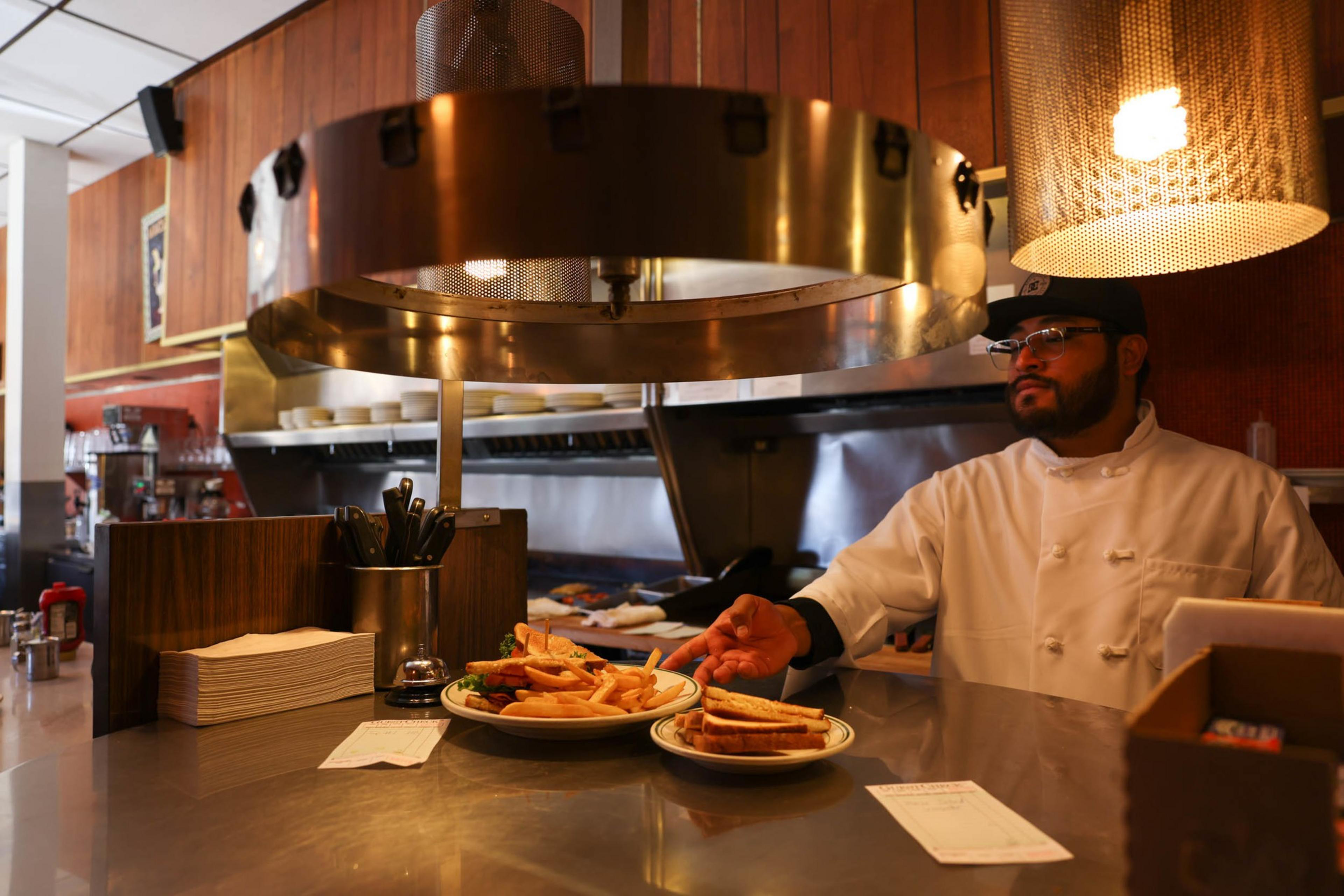 A chef in a white coat and black cap stands behind a counter with plates of fries, sandwiches, utensils, and order tickets in a warmly lit kitchen.