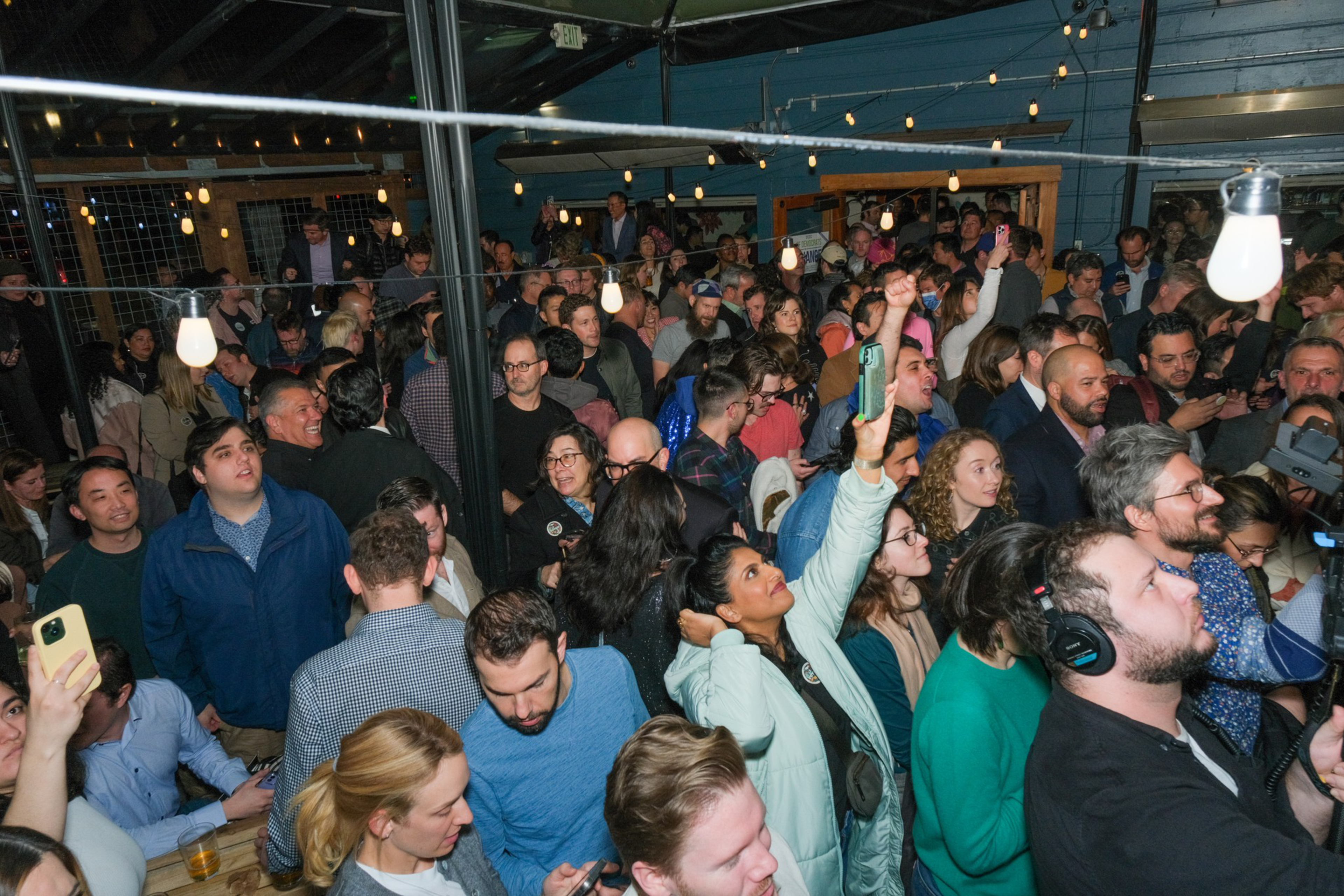 A crowded indoor event with people looking attentively, some holding phones, under string lights at Anina cocktail lounge.