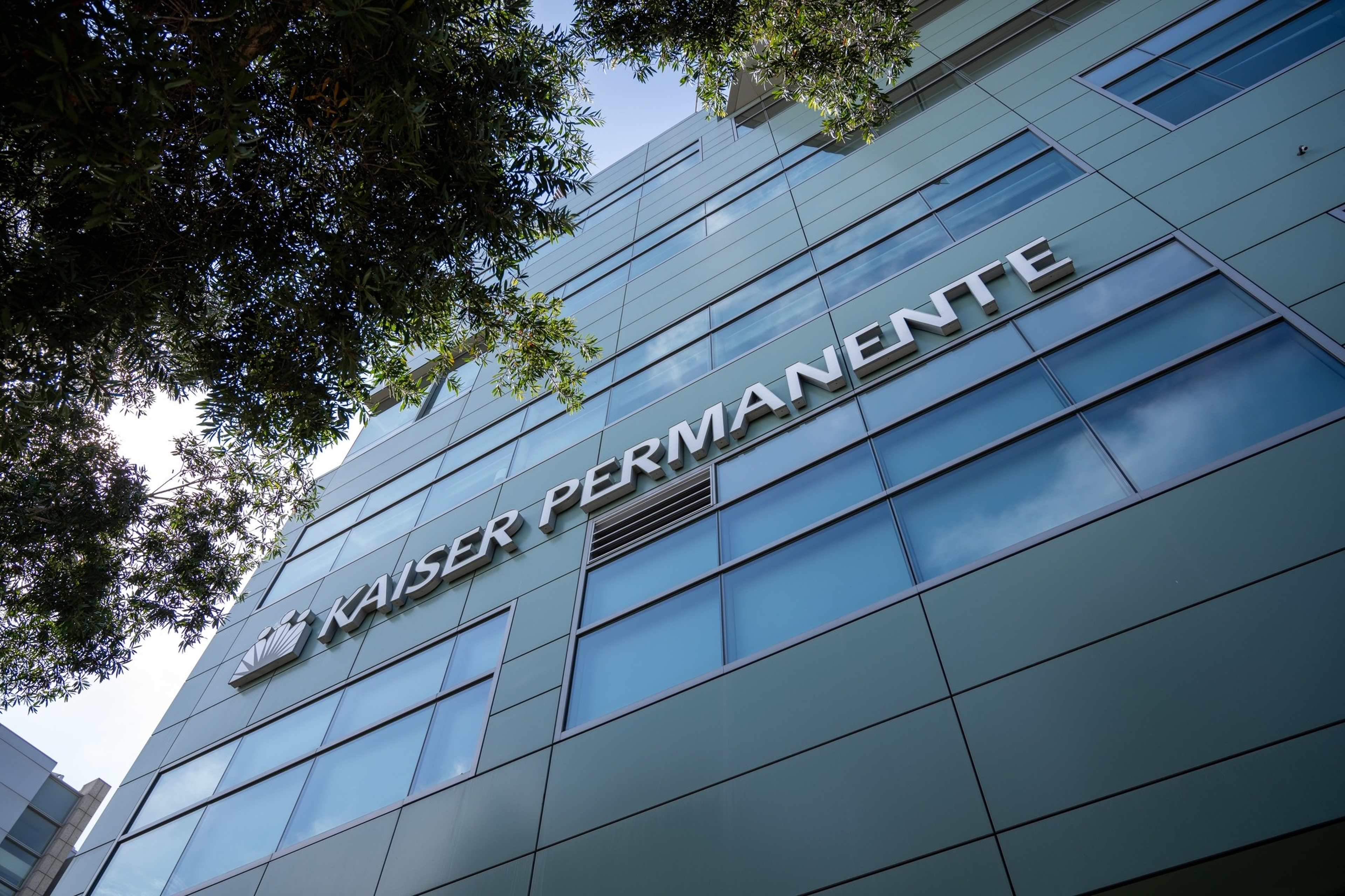 A modern glass and metal building displays the words "KAISER PERMANENTE" near the top, with tree branches framing the left side.