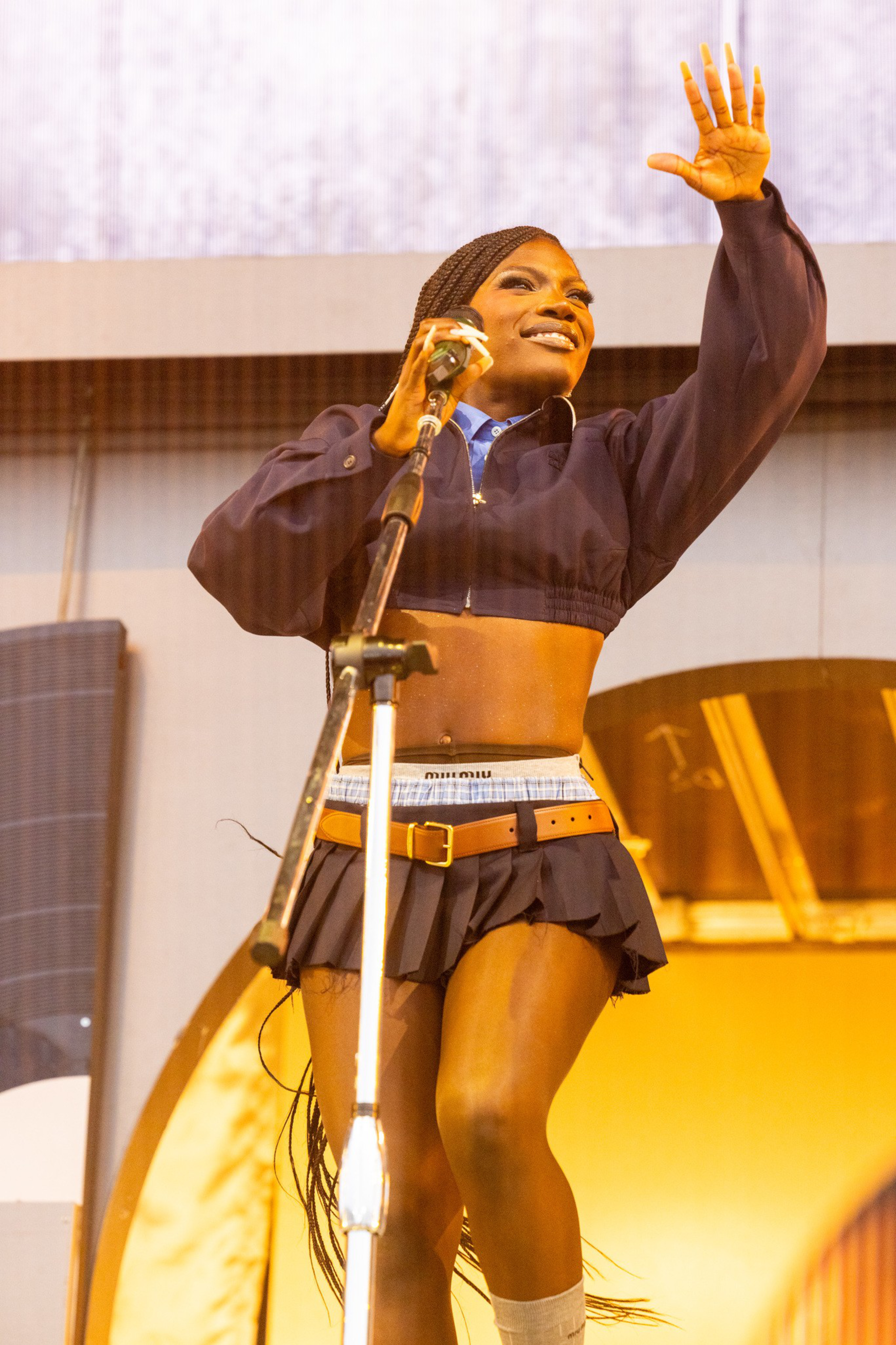 A smiling woman with braided hair sings into a microphone, wearing a cropped jacket, mini skirt, and waving with her free hand on stage.