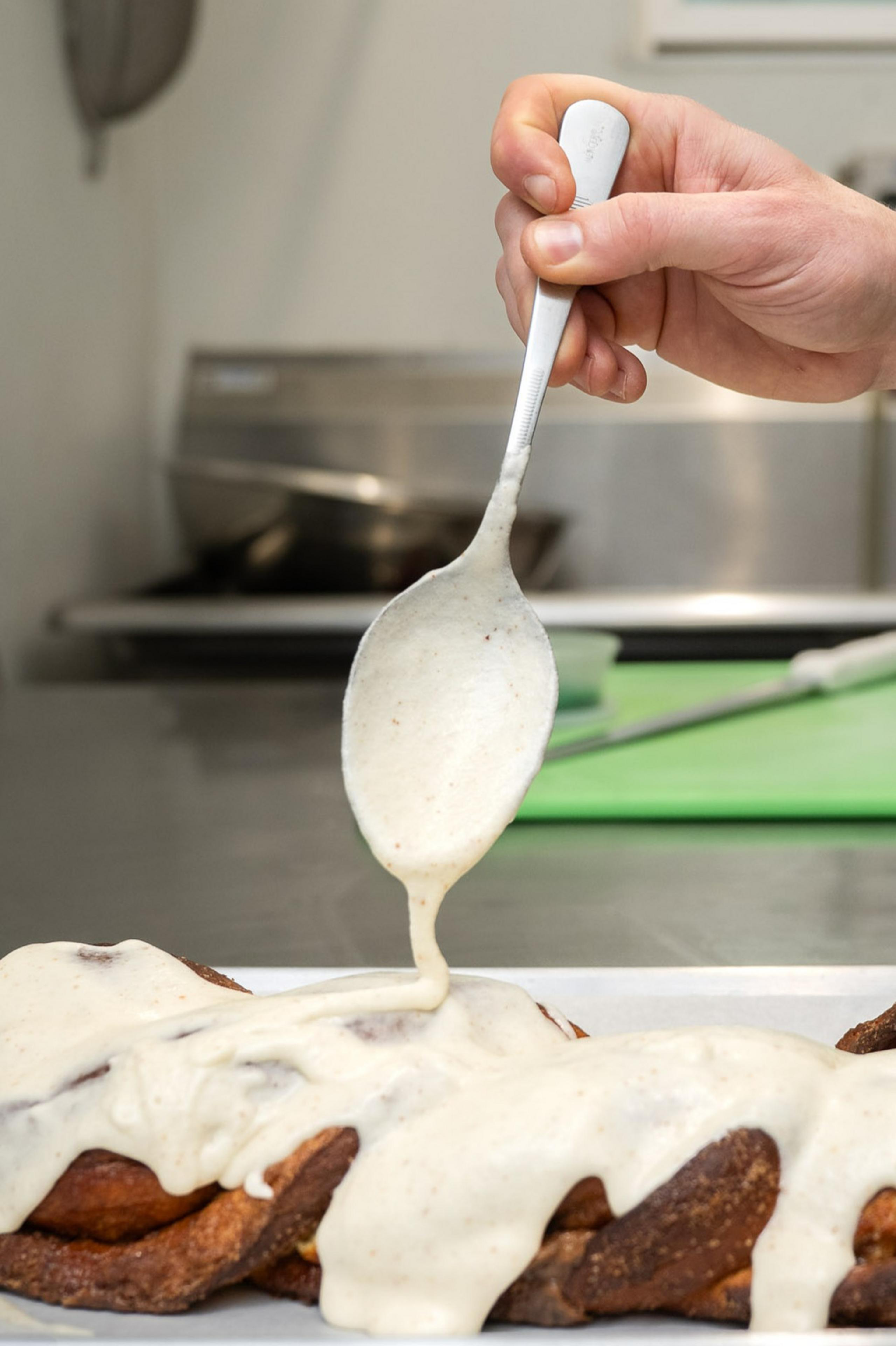 A hand is drizzling creamy icing over a braided pastry with a spoon, in a kitchen setting.