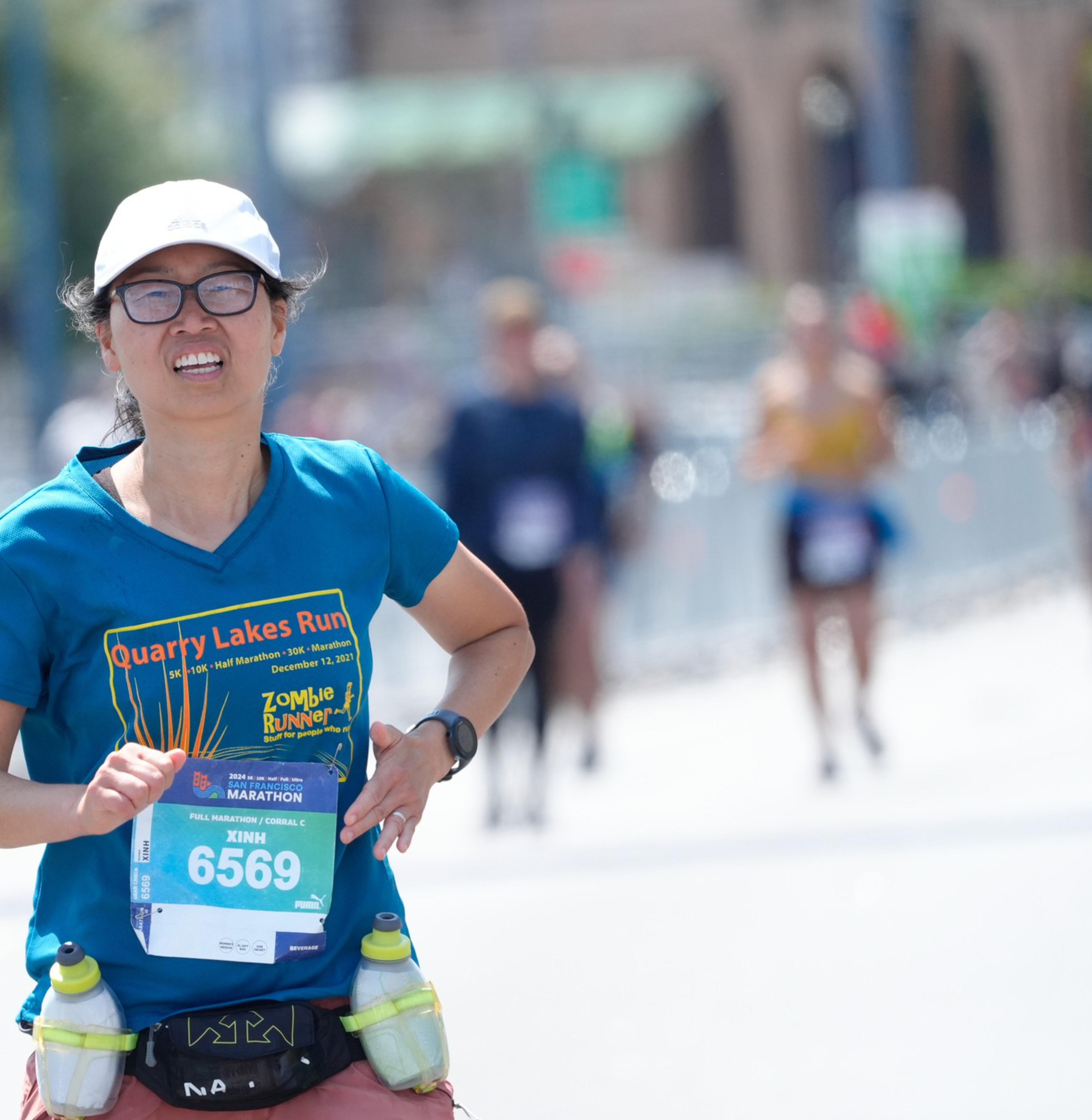 A runner in a blue &quot;Quarry Lakes Run&quot; shirt with race number 6569 is running outdoors, wearing a white cap and glasses, with two water bottles strapped to her waist.