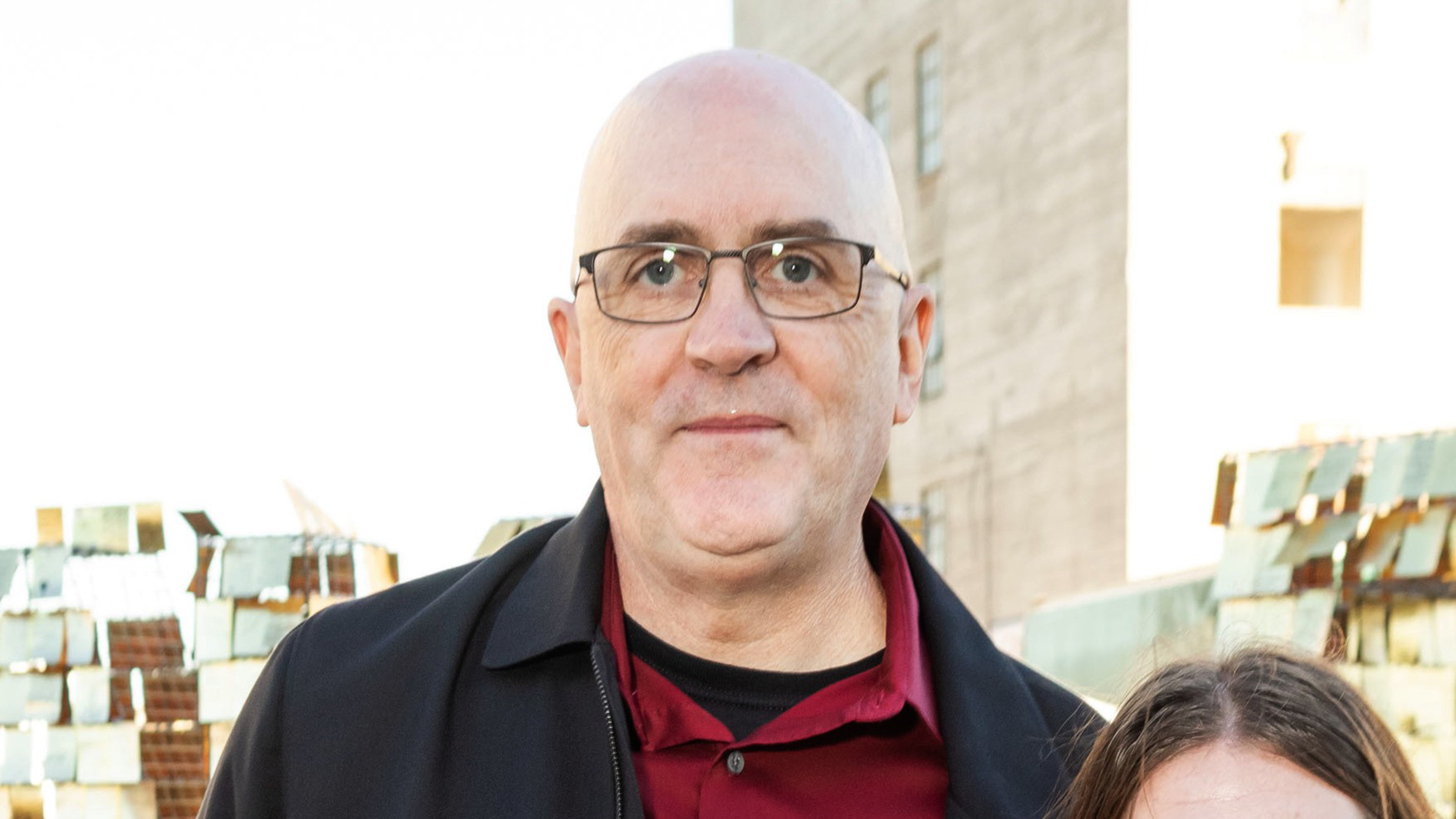 A bald man with glasses is wearing a black jacket over a red shirt, standing outdoors with a building and a metal structure in the background.