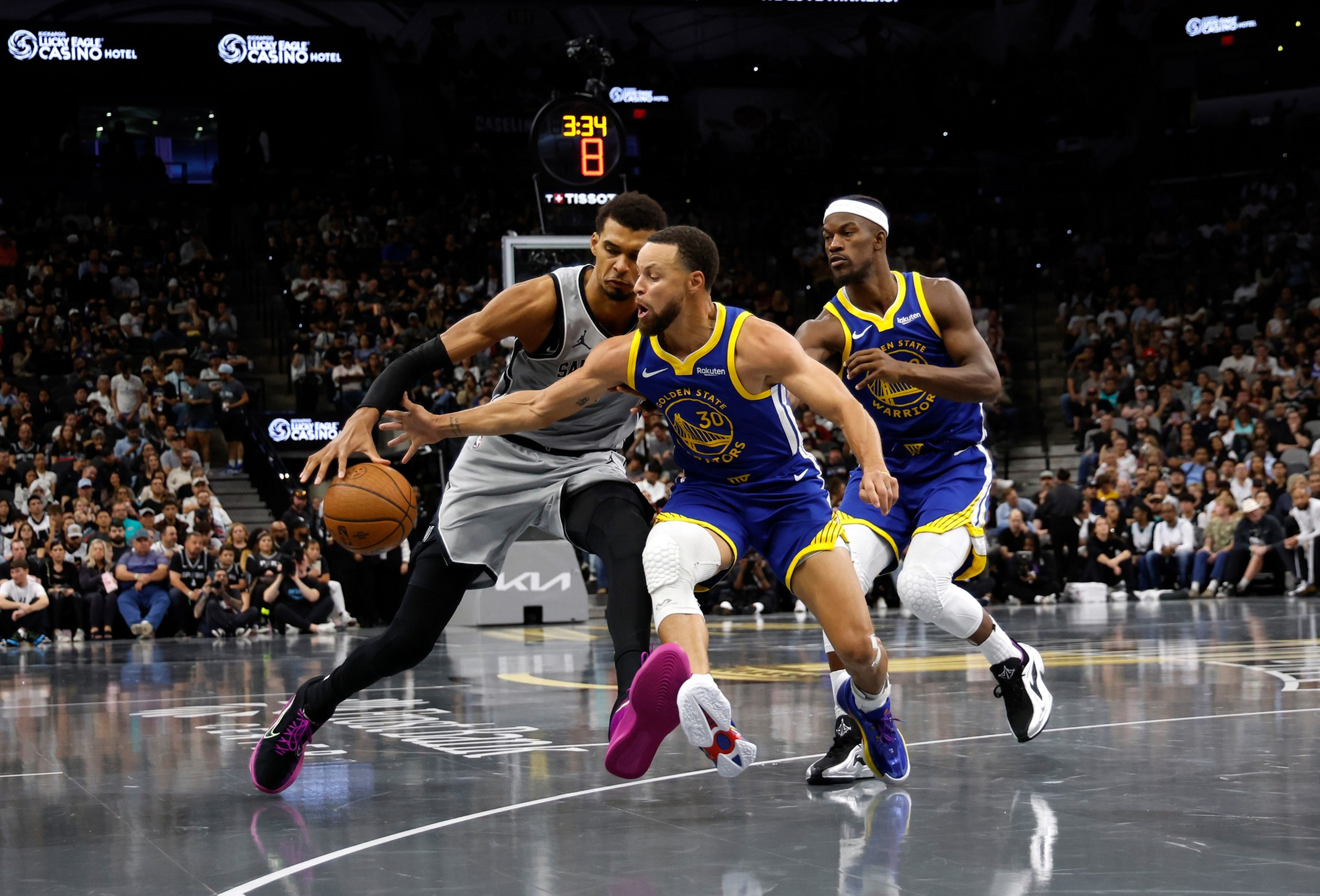 A basketball player in a gray uniform dribbles the ball while being closely guarded by two players in blue and yellow Golden State Warriors uniforms, with a crowd in the background.