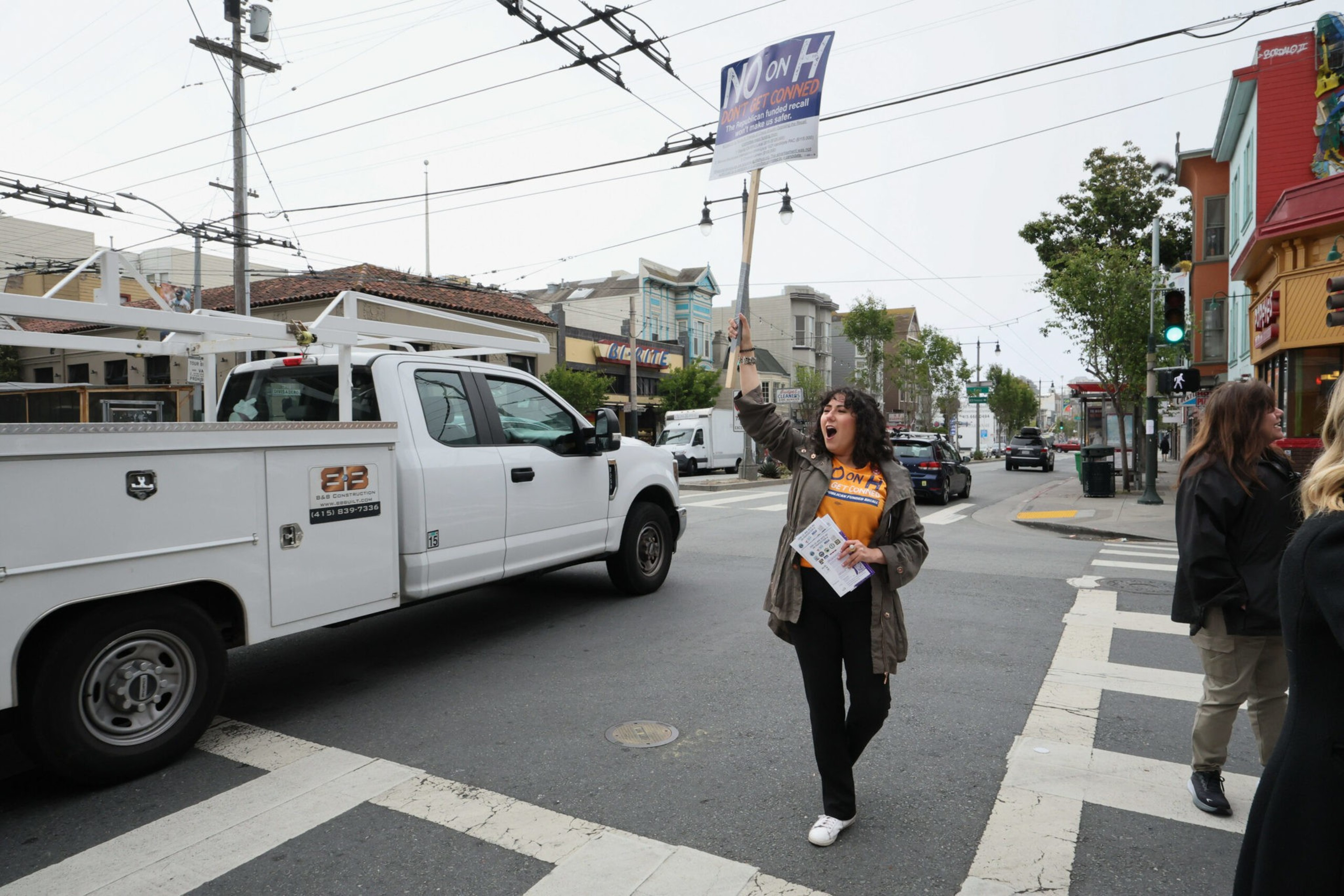 A woman stands at a crosswalk holding a protest sign that reads "No on H," appears to be shouting, with a white truck passing nearby.