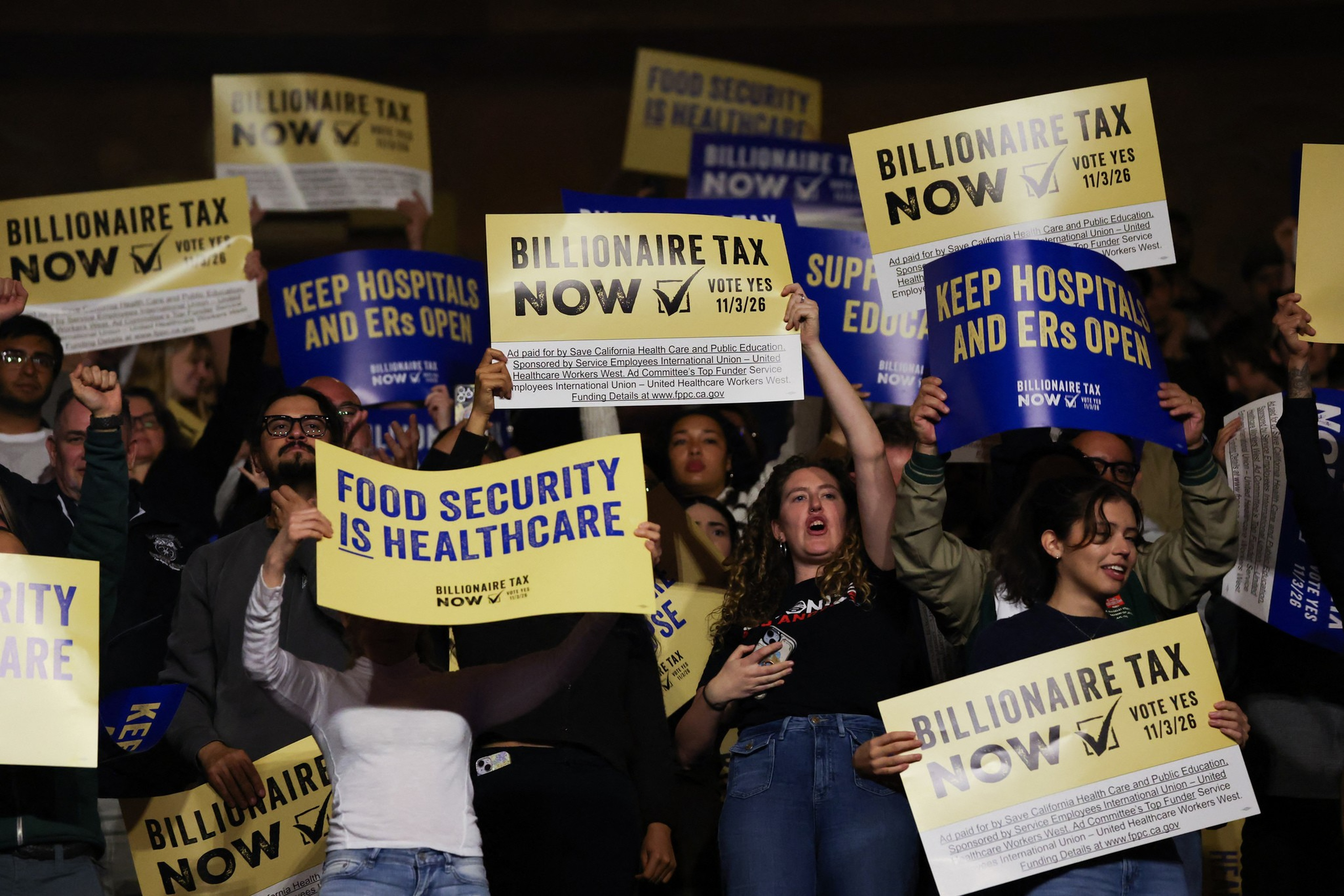 A diverse group of people hold signs advocating for “Billionaire Tax Now,” “Food Security is Healthcare,” and “Keep Hospitals and ERs Open” during a rally.