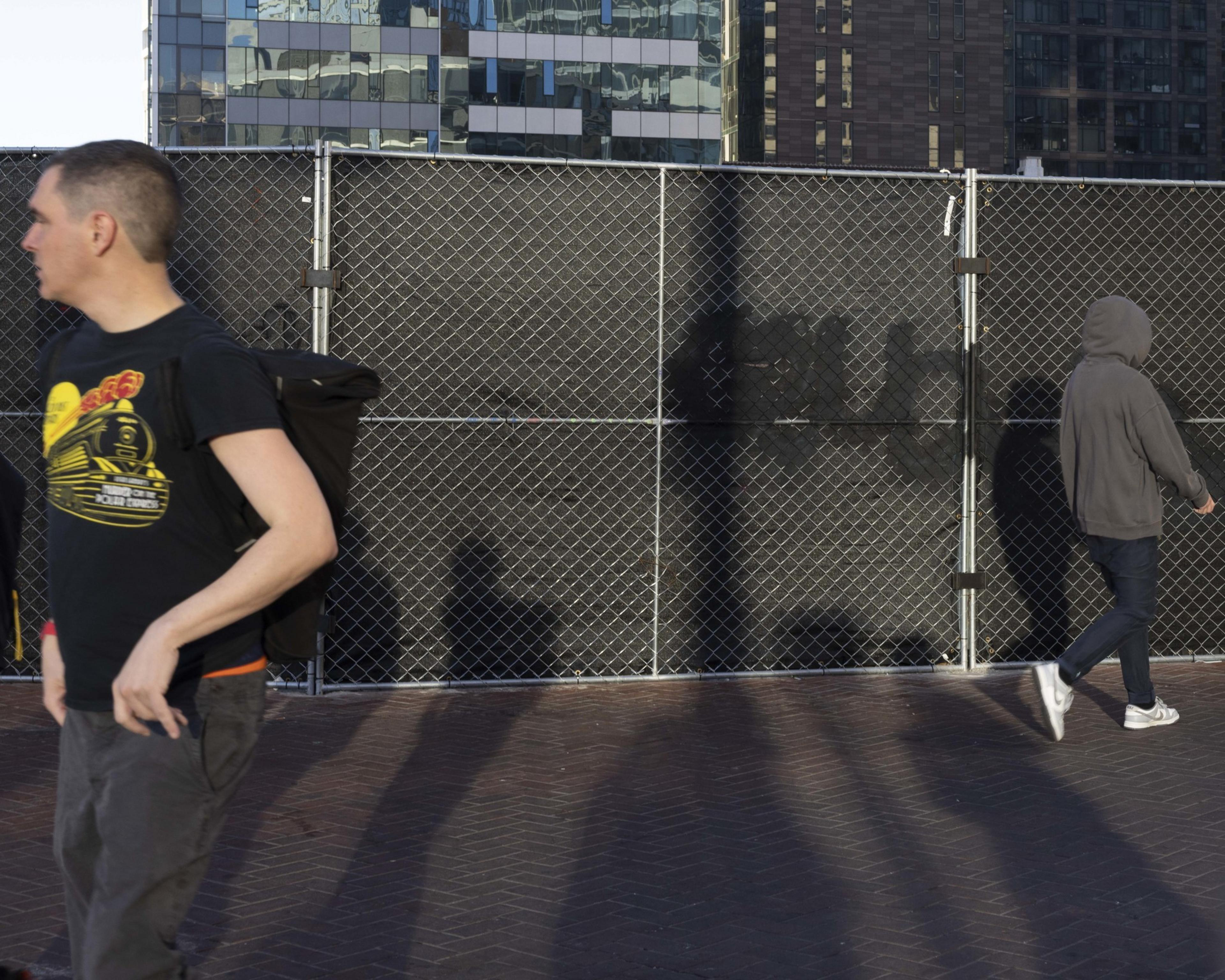 Two people are walking in front of a tall chain-link fence with black privacy fabric; one wears a dark T-shirt and backpack, the other is in a gray hoodie, jeans, and sneakers.