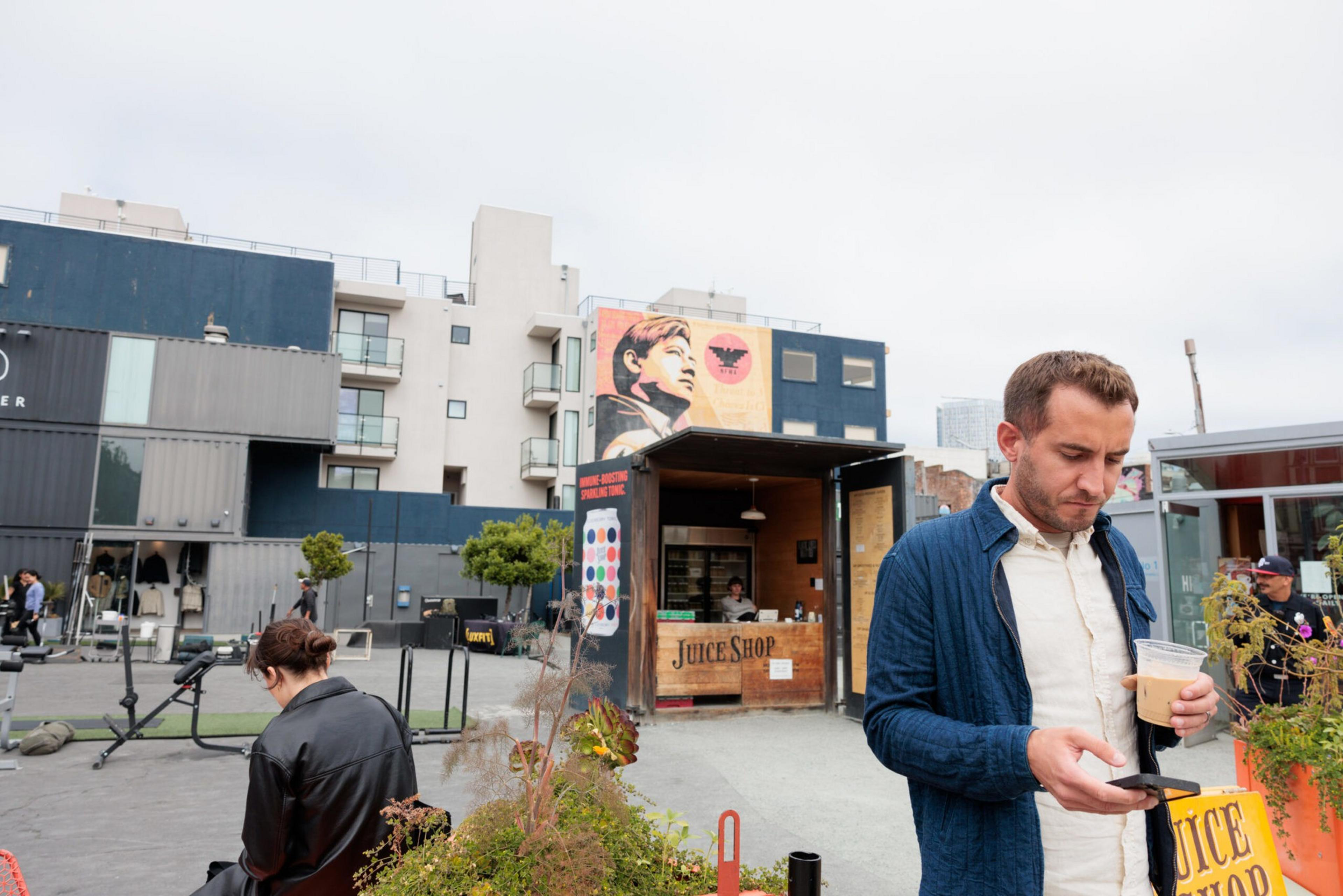 A man in a blue jacket drinks coffee and looks at his phone near a small juice shop kiosk, with a mural and apartment buildings in the background.