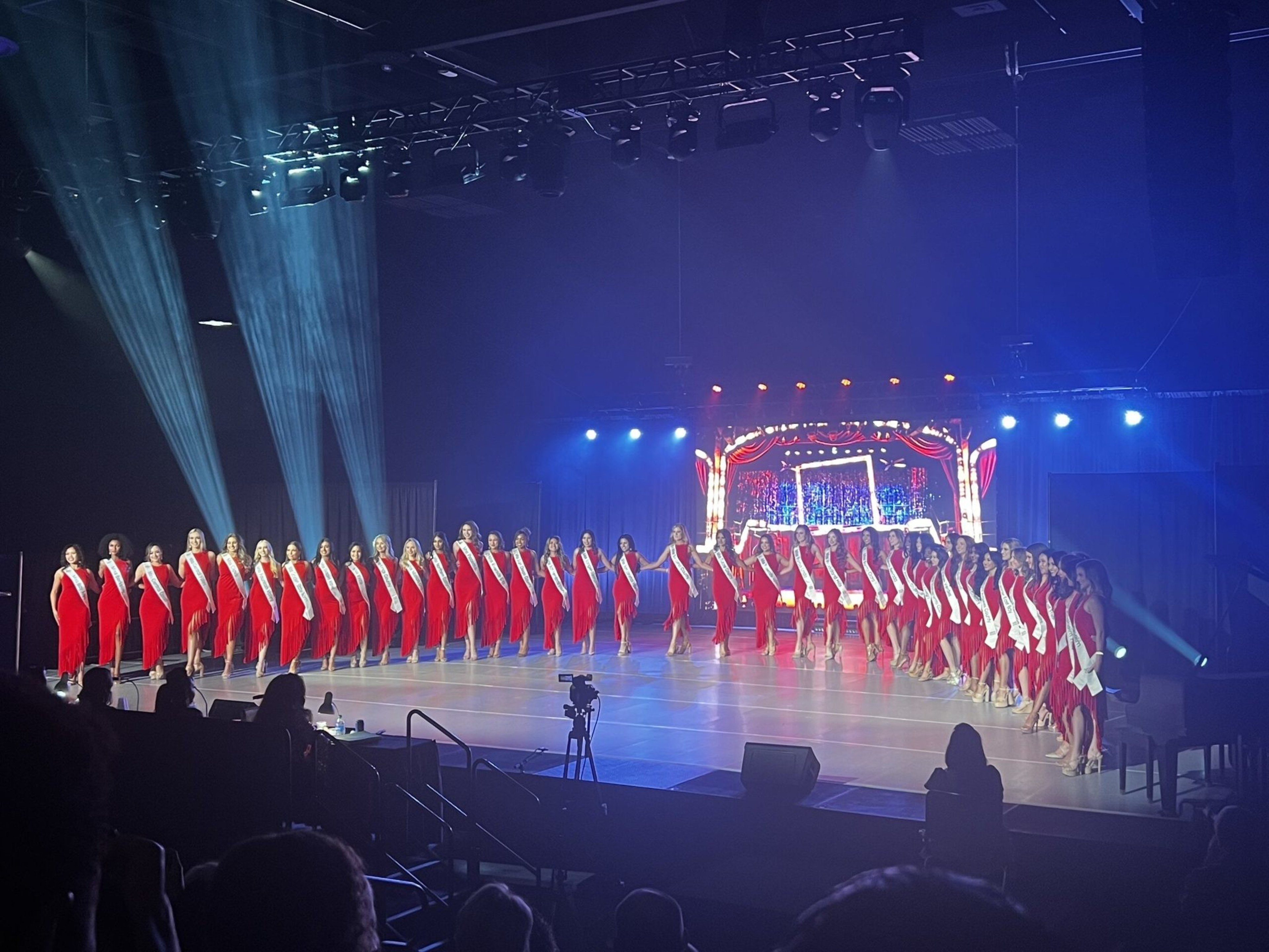 Women wearing red dresses and white sashes stand in a large curved line on stage, illuminated by blue and white stage lights with a decorative backdrop.