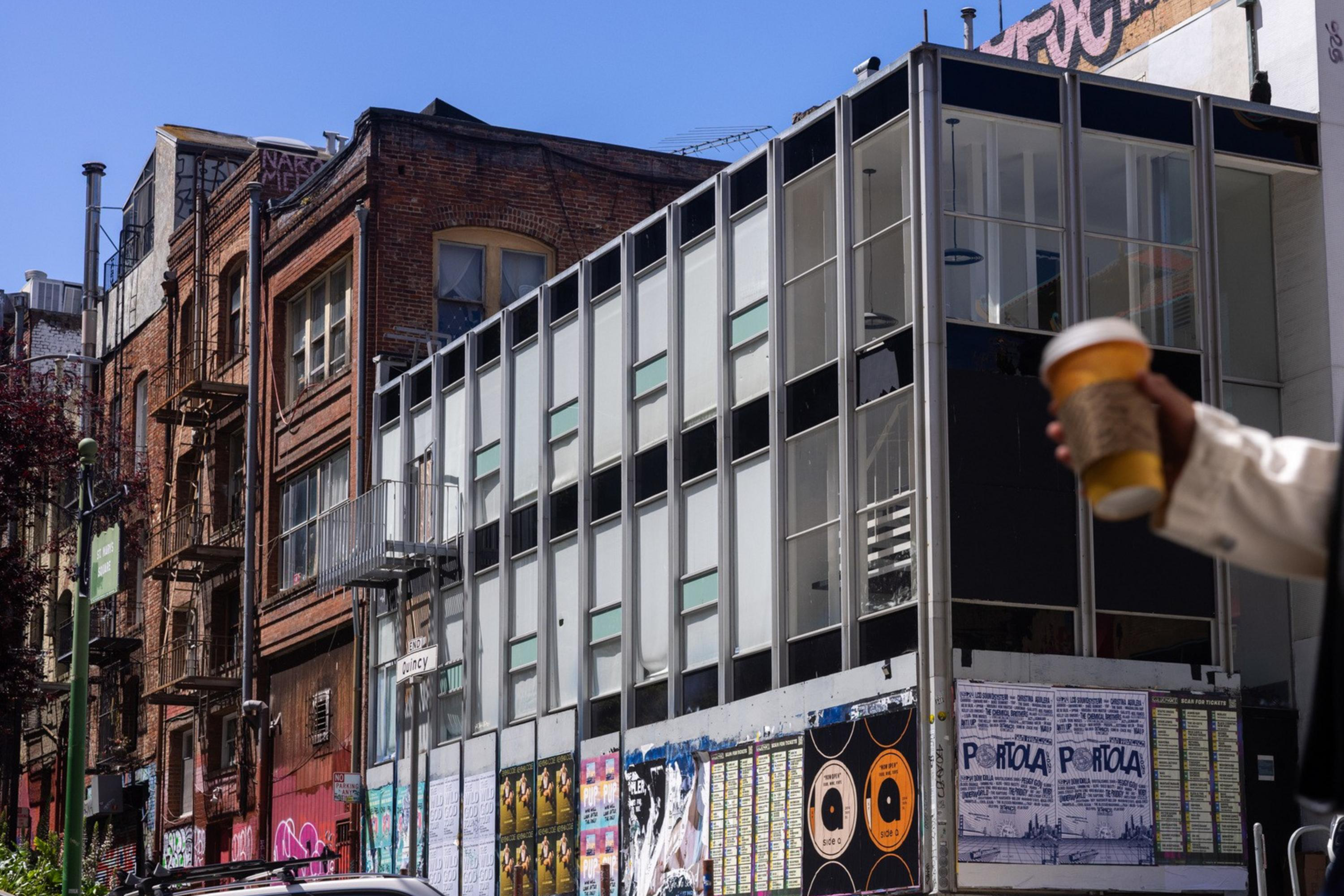 A woman holding a coffee cup walks past a modern glass building with posters on it. Old brick buildings line the street, and a white car is parked nearby.