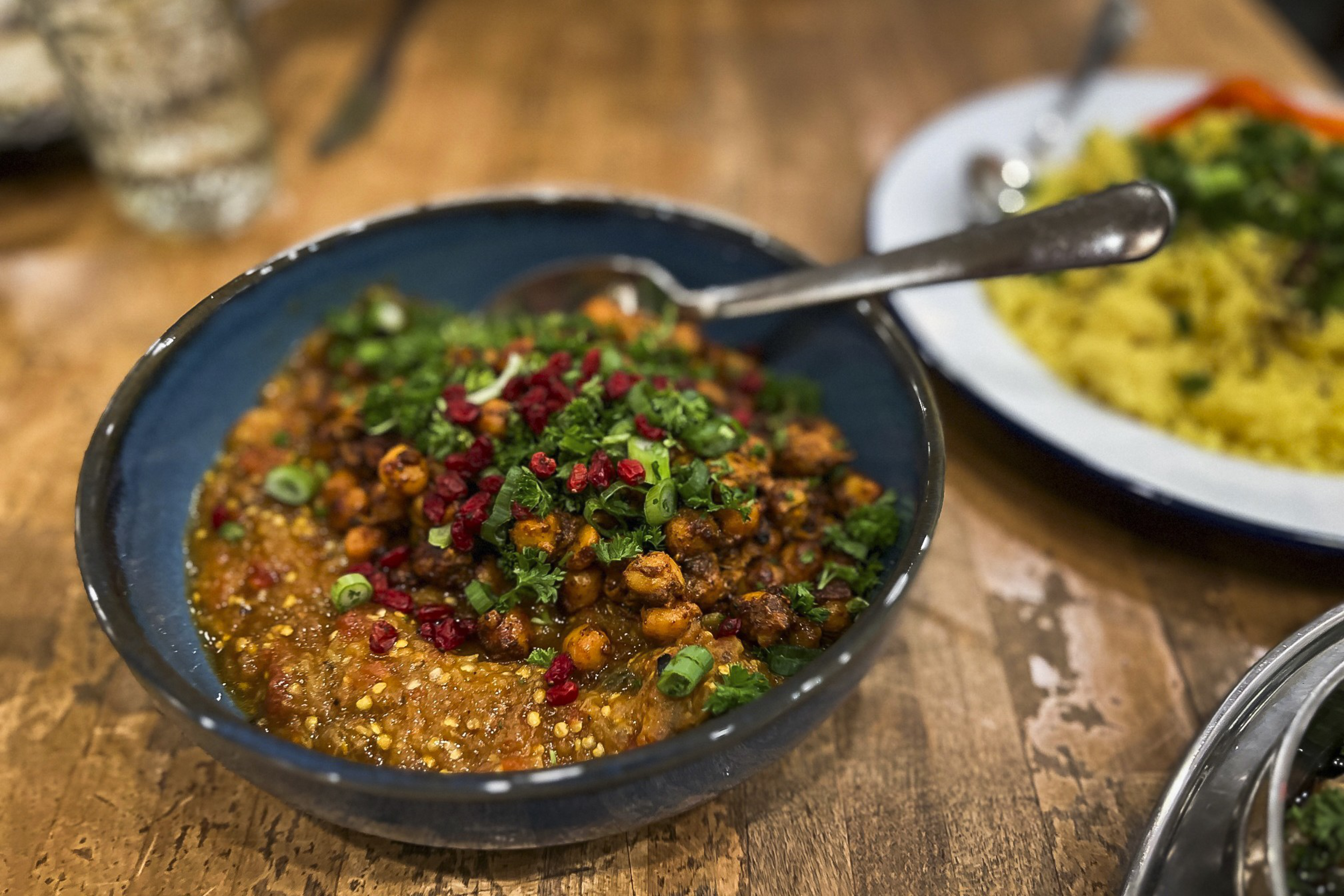 A bowl of spicy chickpea dish garnished with green herbs and red bits, with other plates of food on a wooden table.