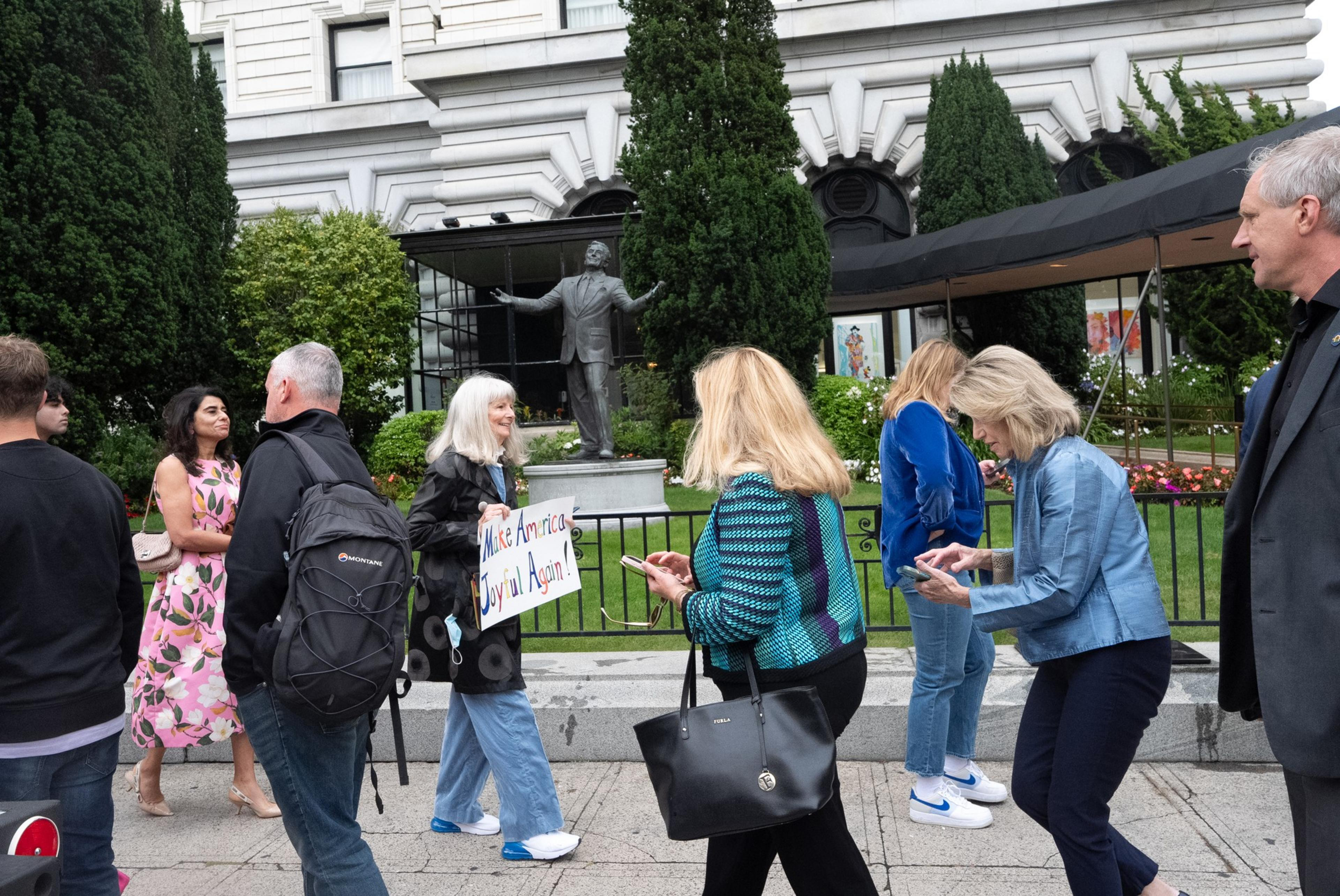 A group of people walk in front of a building, one holding a sign that reads, &quot;Make America Joyful Again!&quot; A statue with outstretched arms stands in the background.