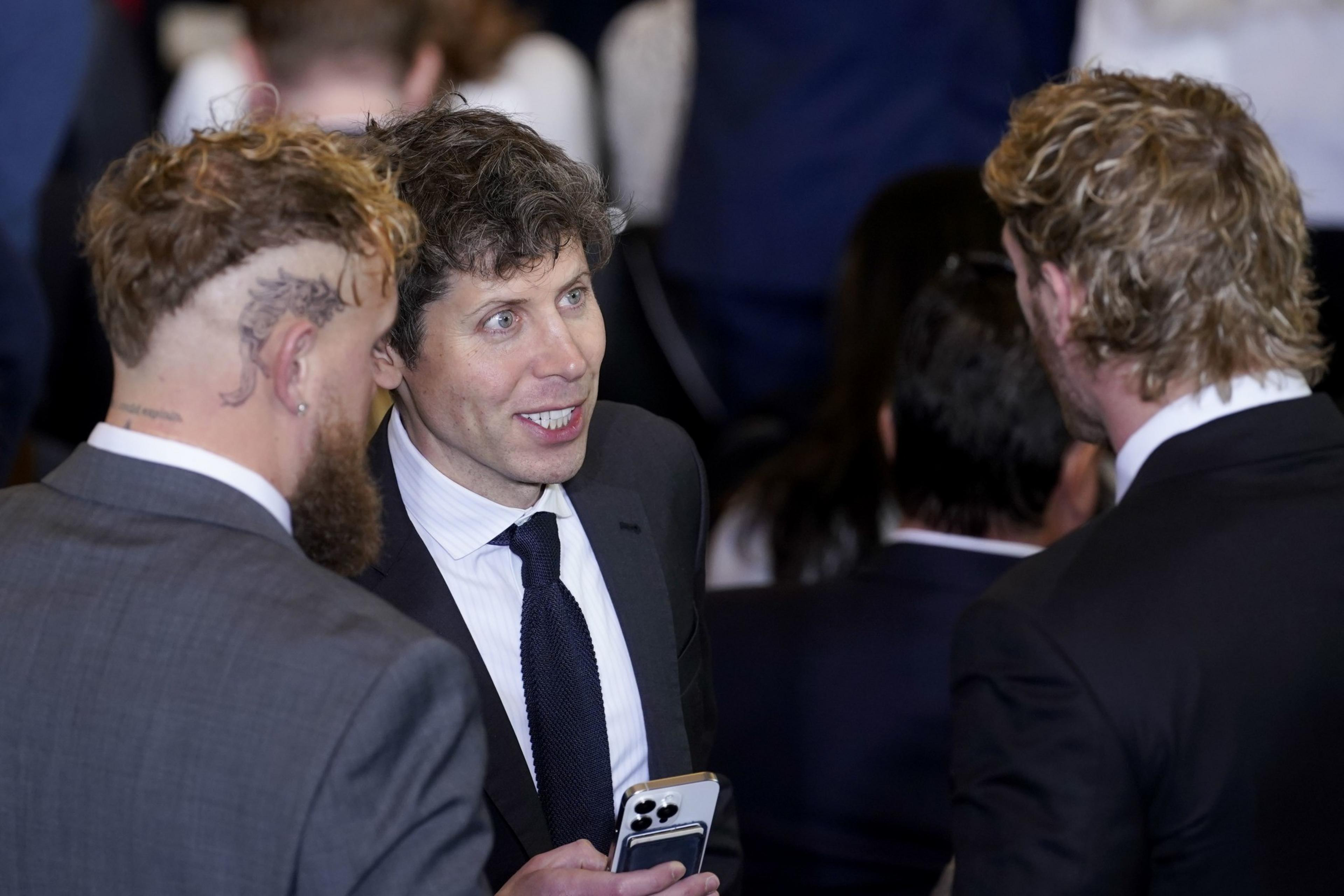 Three men in suits are engaged in conversation. One has a tattoo on his head, another holds a phone. They appear to be in a crowded social setting.