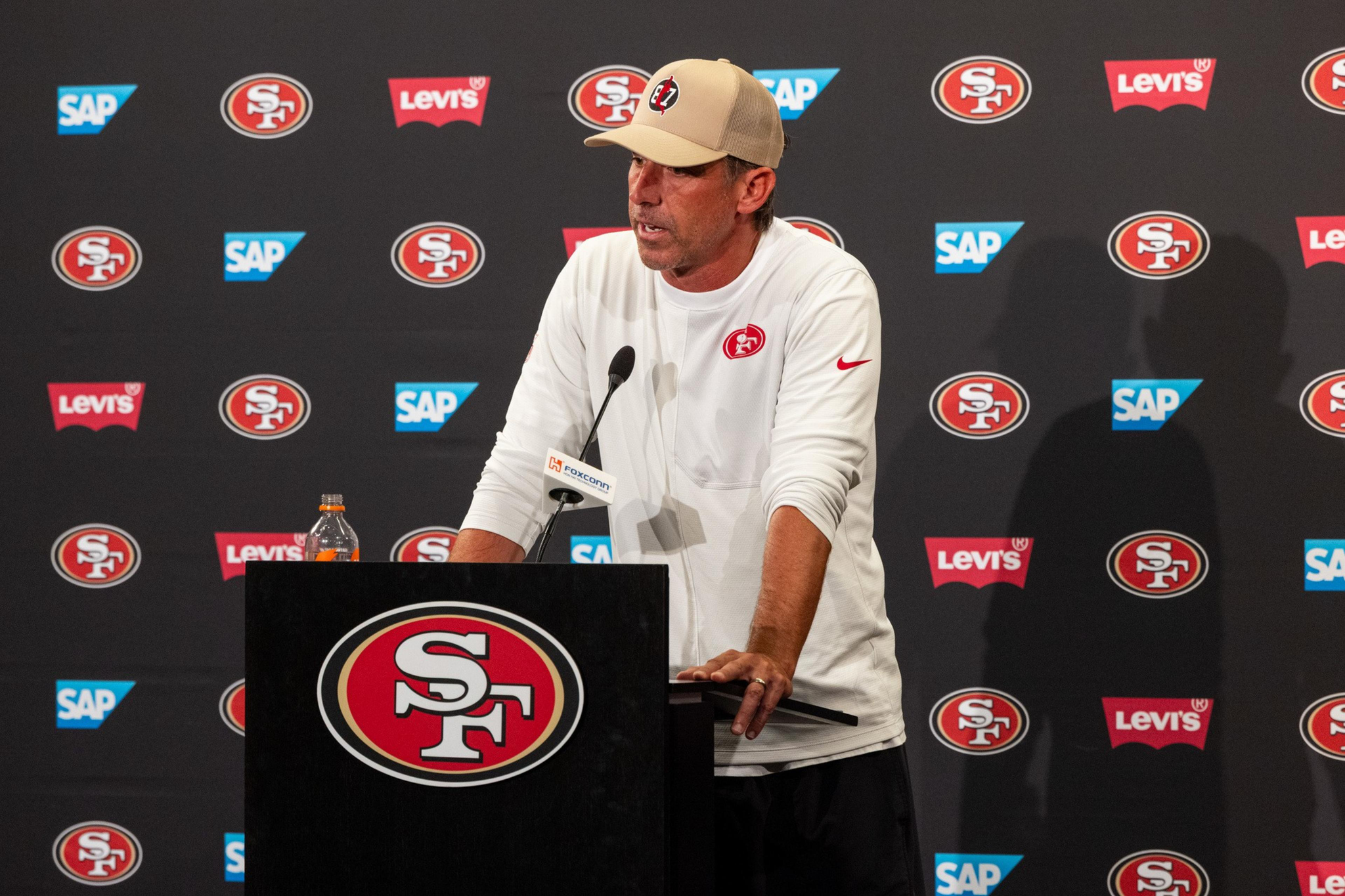 A man in a beige cap and white shirt with a red 49ers logo speaks at a podium covered with the 49ers emblem, backed by a wall of sponsor logos.