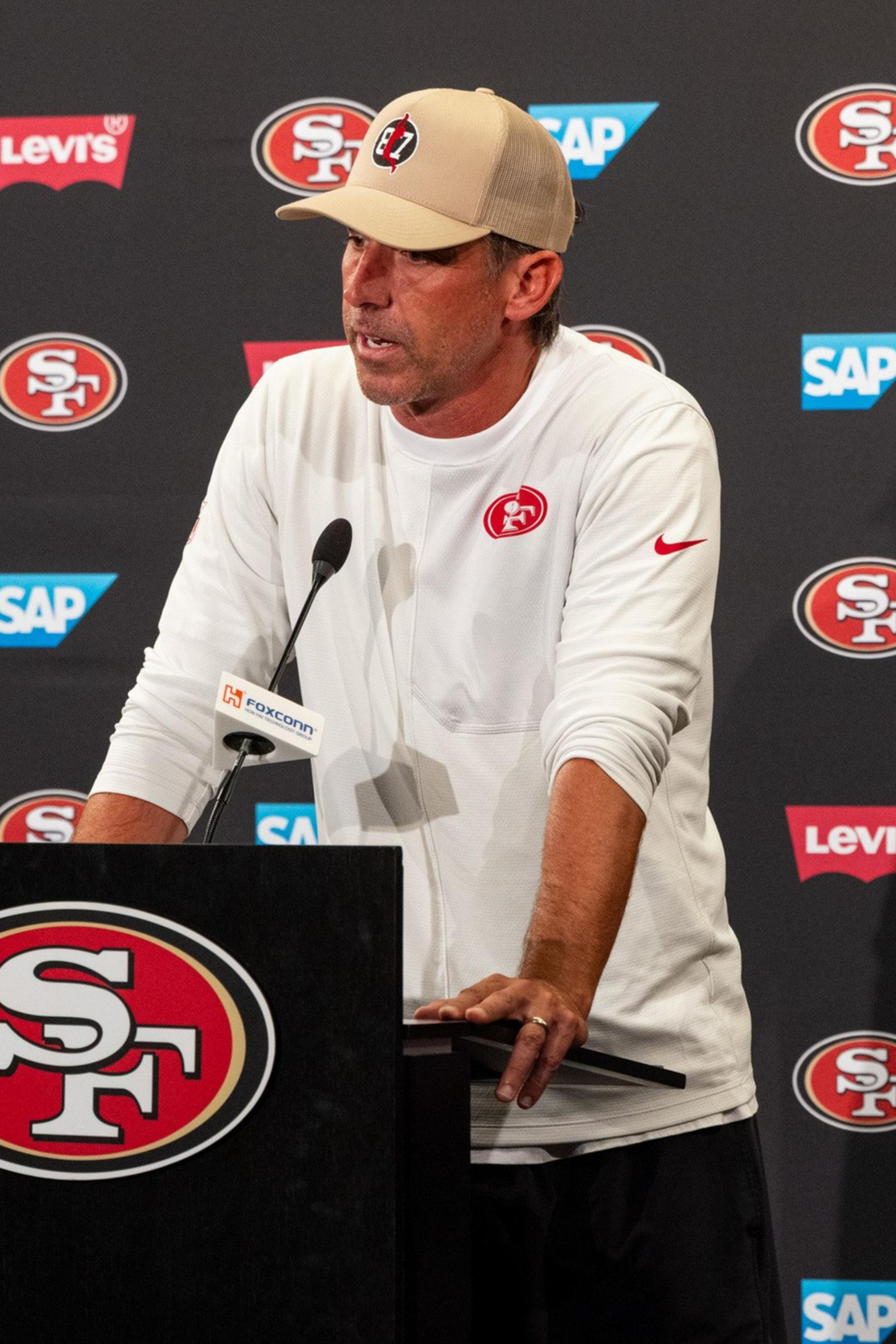 A man in a beige cap and white shirt with a red 49ers logo speaks at a podium covered with the 49ers emblem, backed by a wall of sponsor logos.