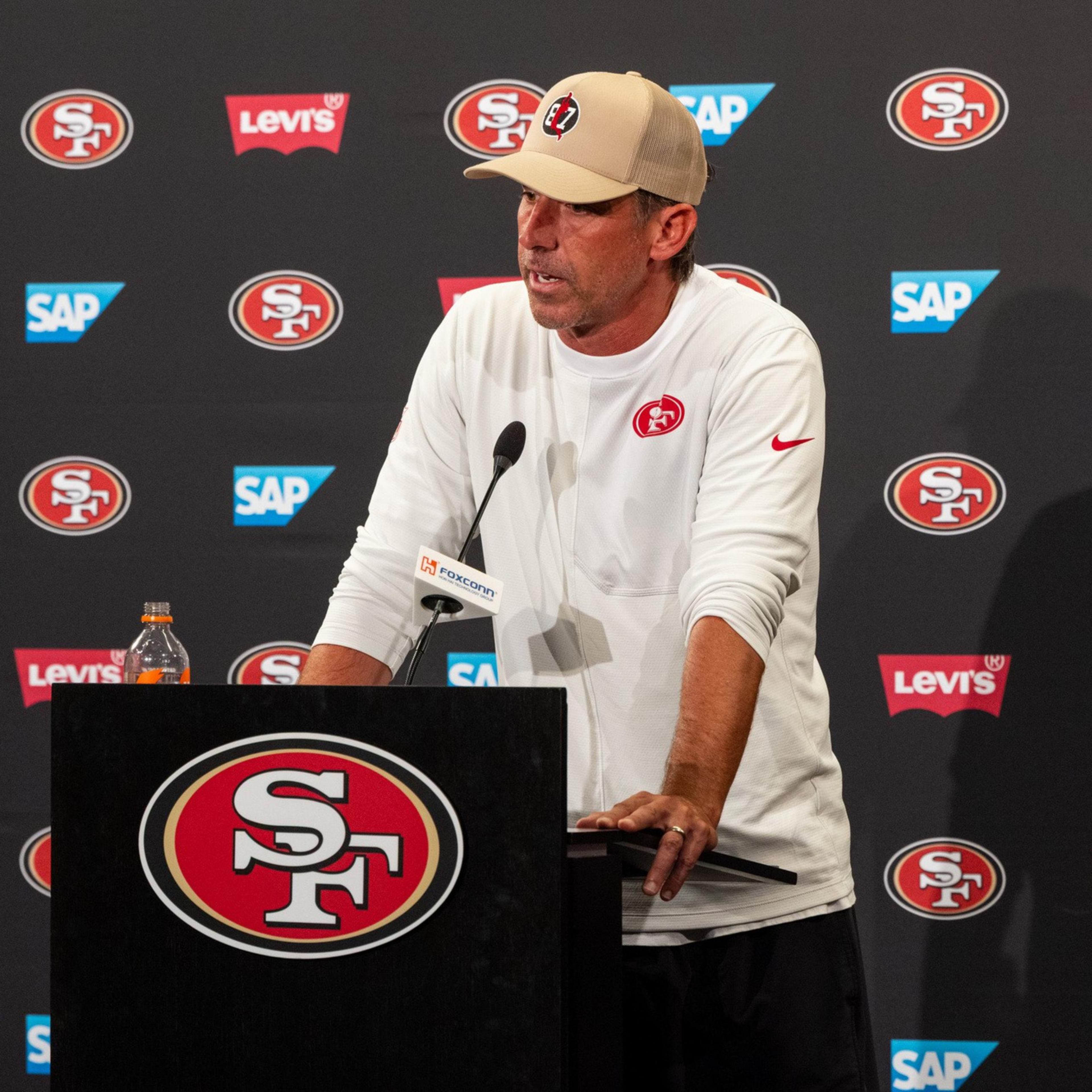 A man in a beige cap and white shirt with a red 49ers logo speaks at a podium covered with the 49ers emblem, backed by a wall of sponsor logos.
