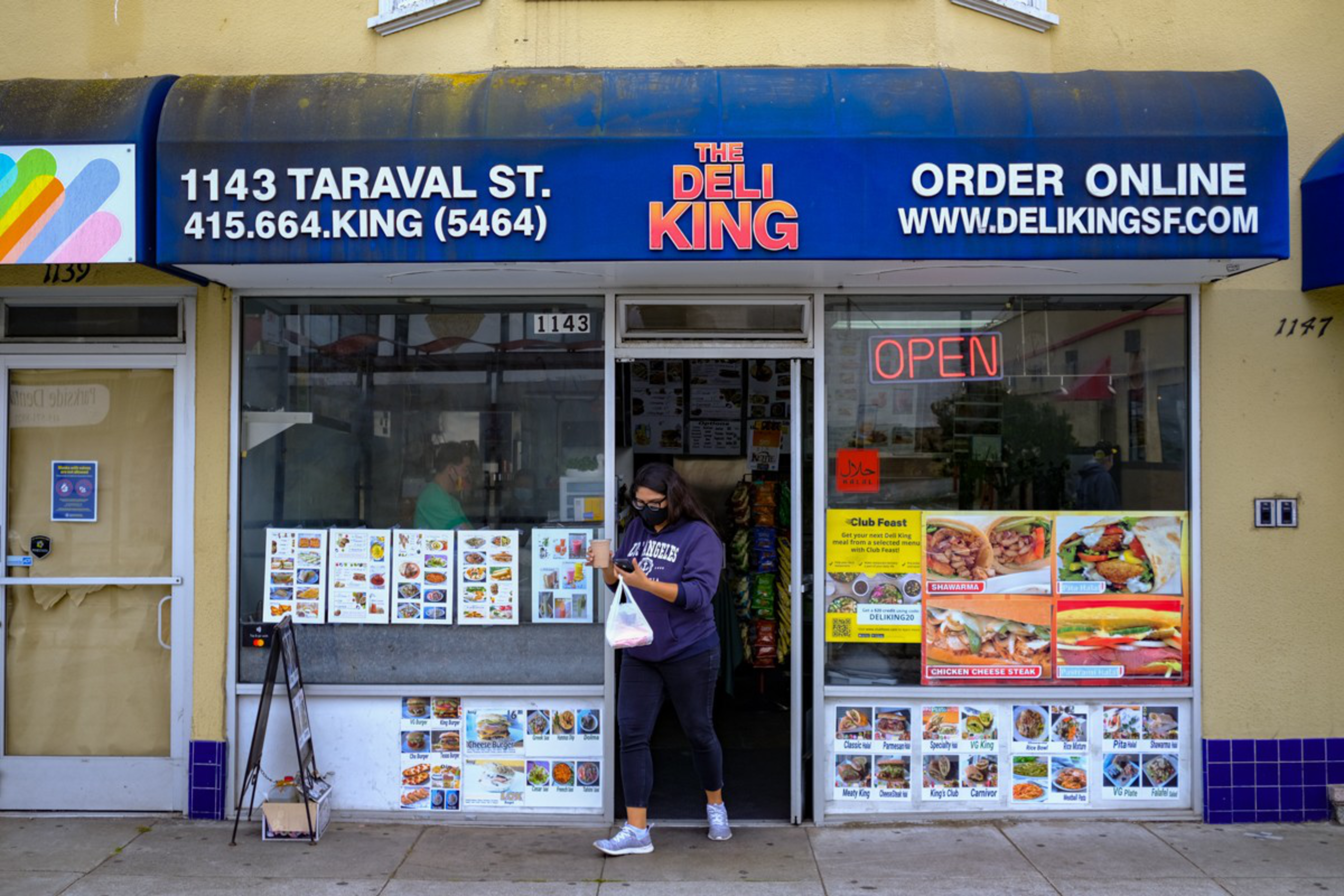 A woman wearing a mask exits The Deli King at 1143 Taraval St., holding a takeout bag, with menu images and a neon "OPEN" sign visible in the windows.