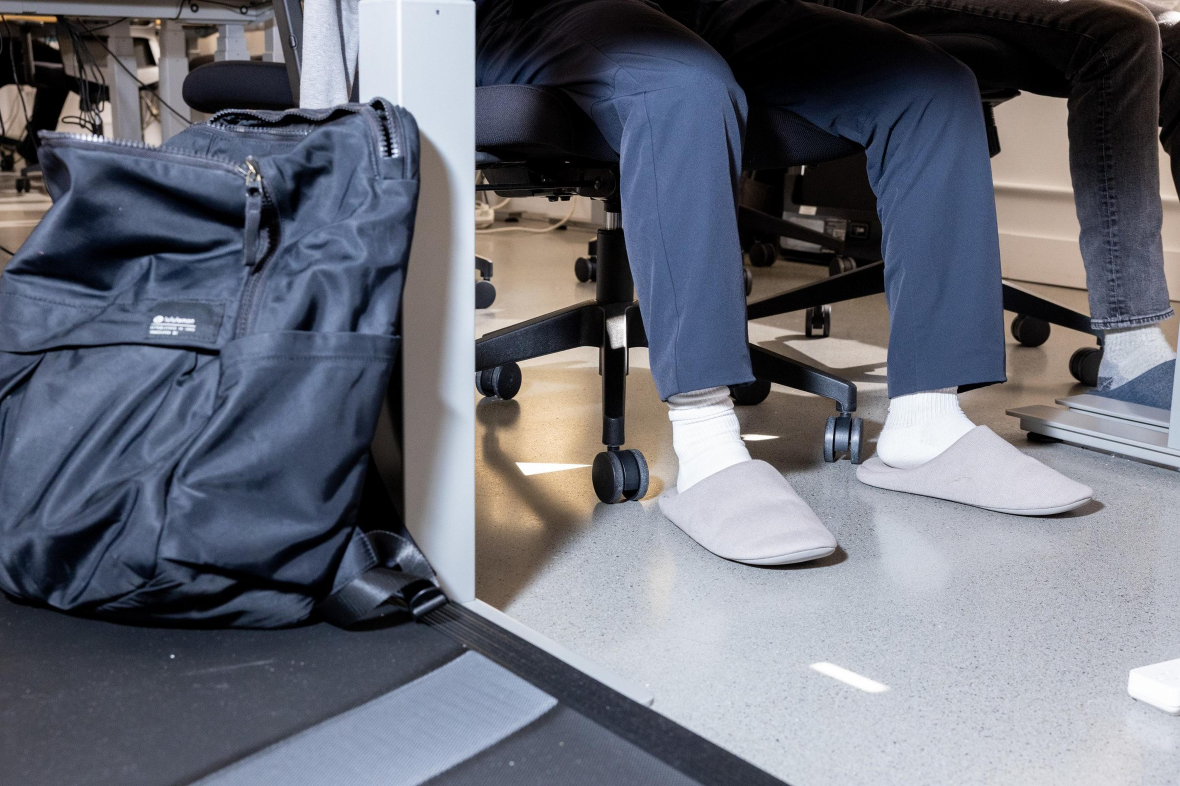 Two people sit on office chairs wearing slippers; one person has white socks and grey slippers, and a black backpack rests nearby on the floor.