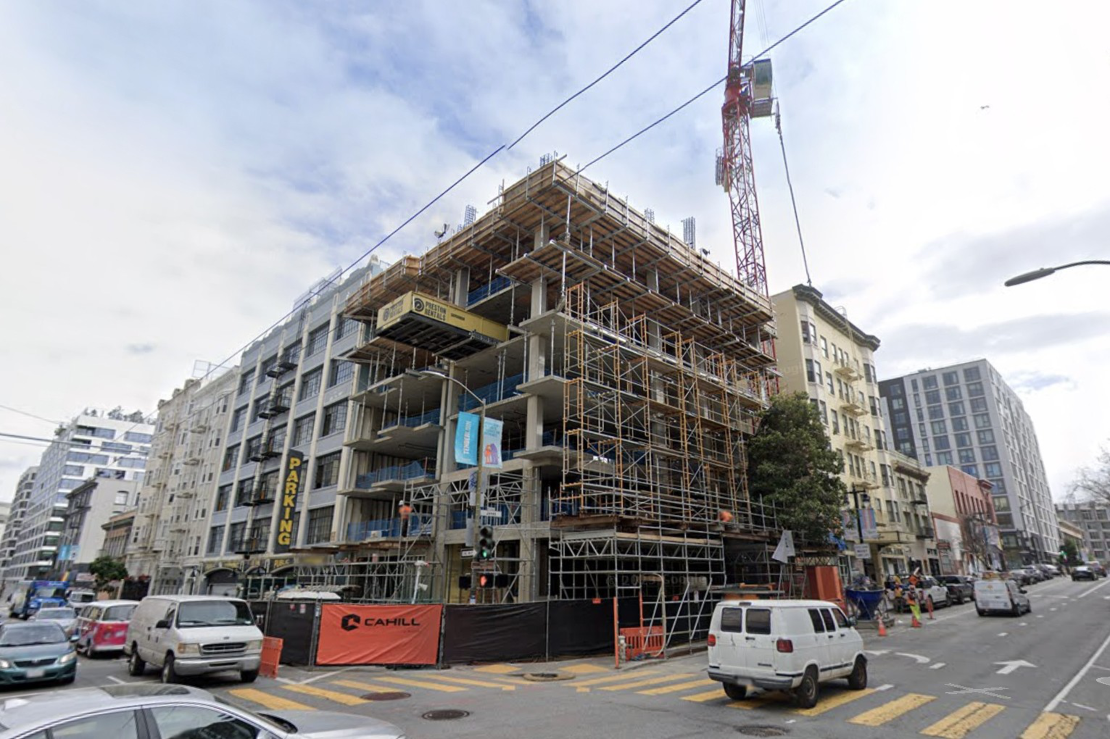 A multi-story building under construction with scaffolding and a crane, surrounded by parked vehicles and adjacent older buildings on a city street.