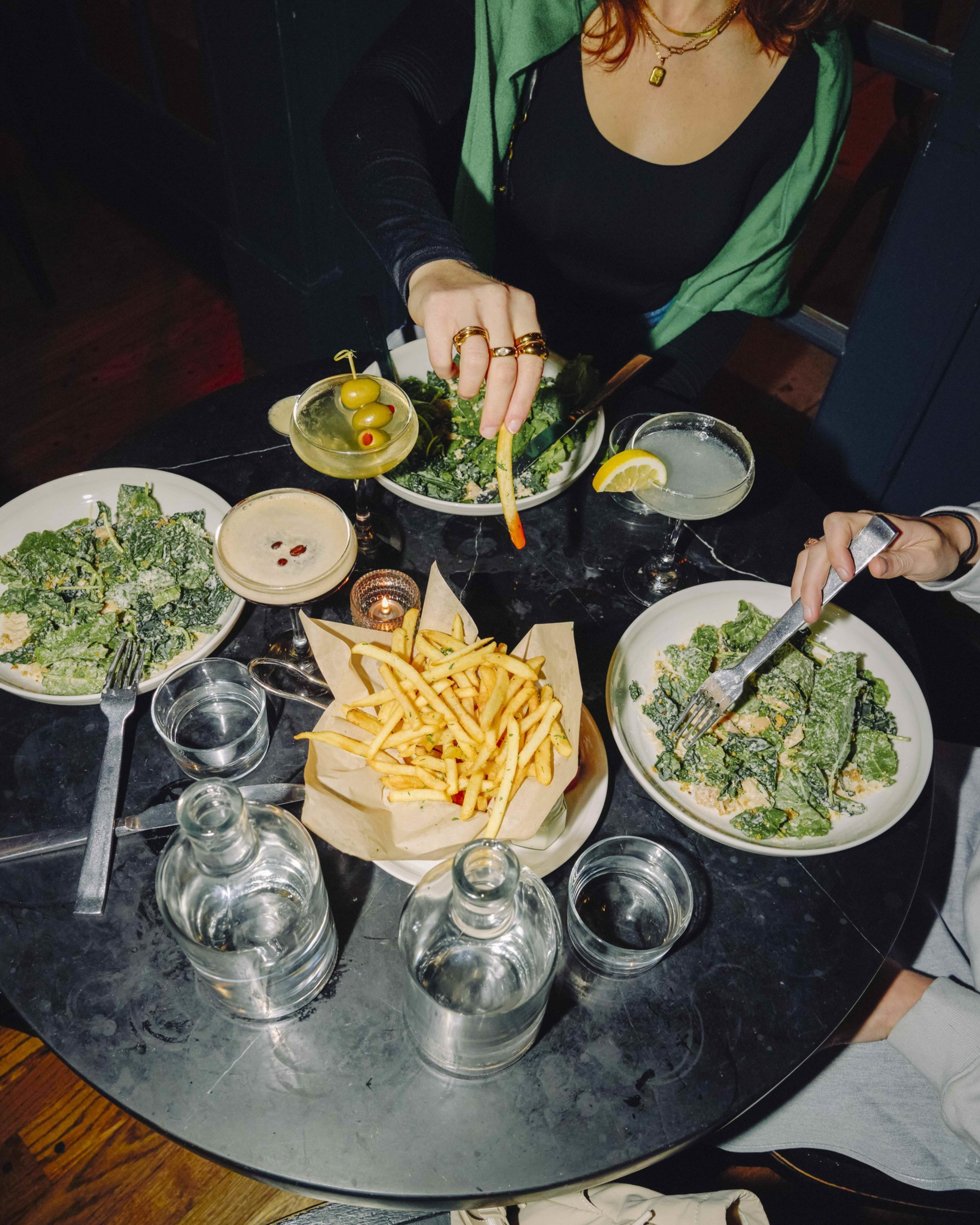 Two people are seated at a table with plates of Caesar salad, a basket of French fries, cocktails, and water bottles, one person is grabbing a fry.