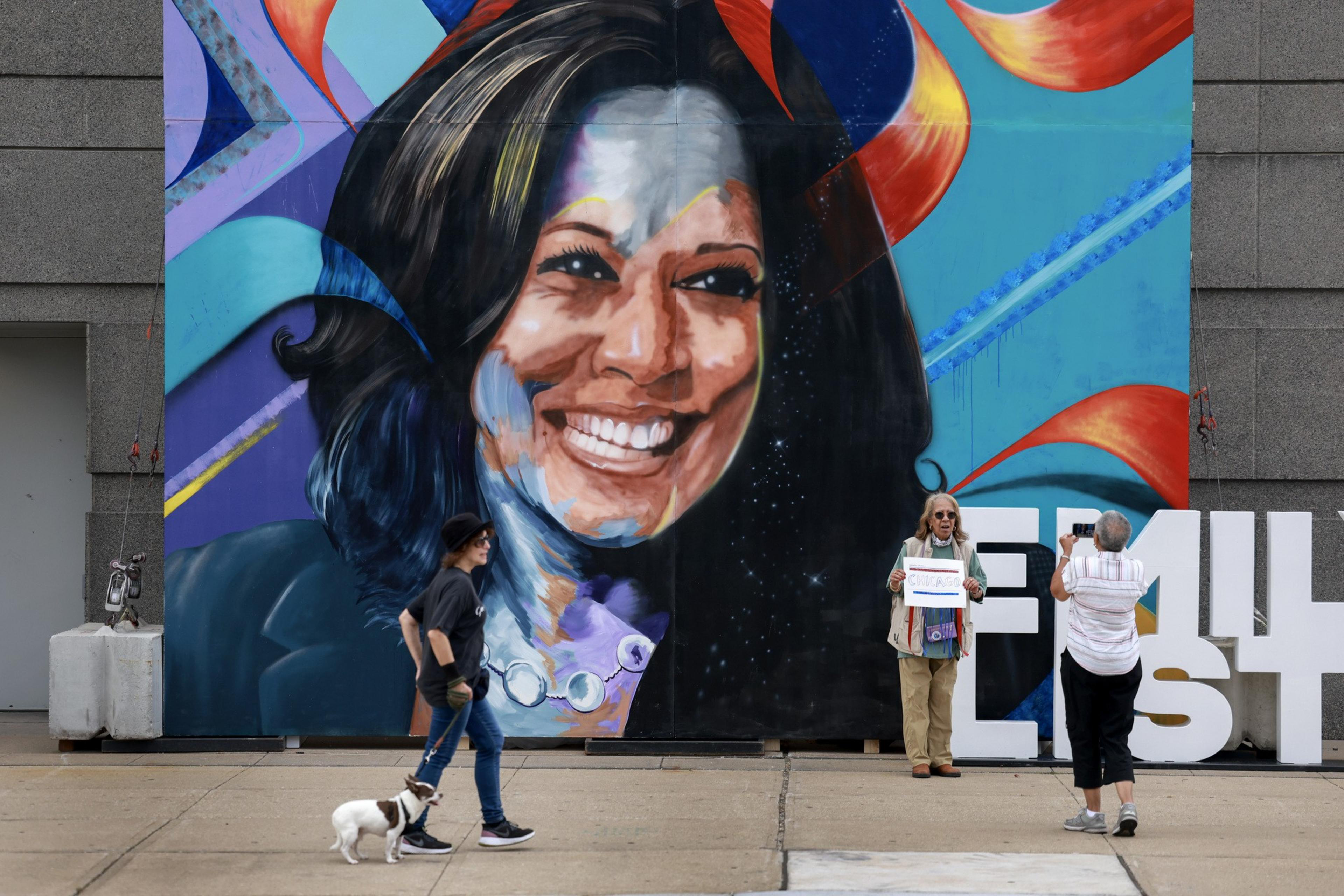The image shows a large, colorful mural of a smiling woman's face on a wall. In the foreground, a person walks a small dog, and two people take a photo near large white letters.