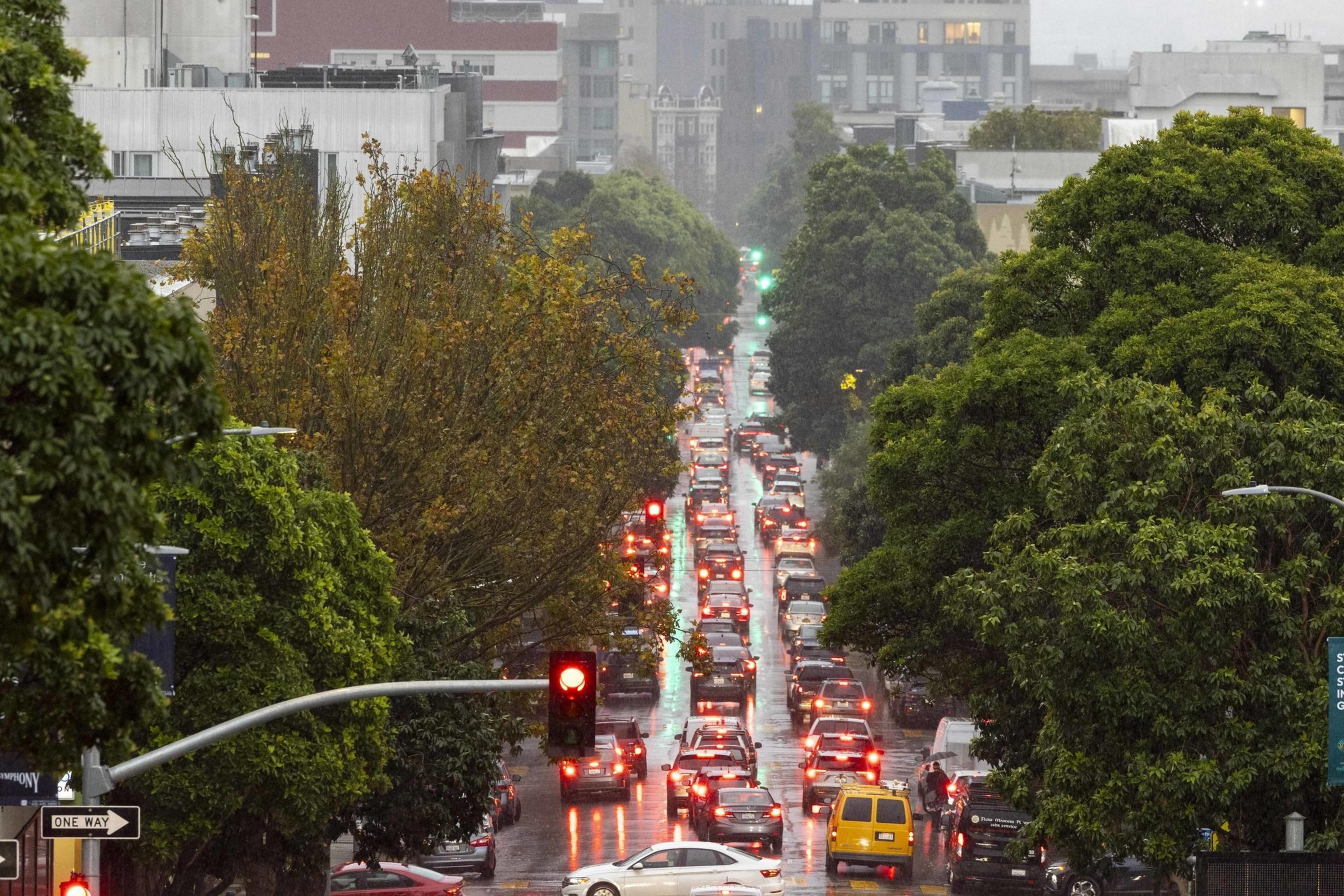 A rainy urban street densely packed with cars and glowing brake lights stretches between lush green trees and buildings under a cloudy sky.