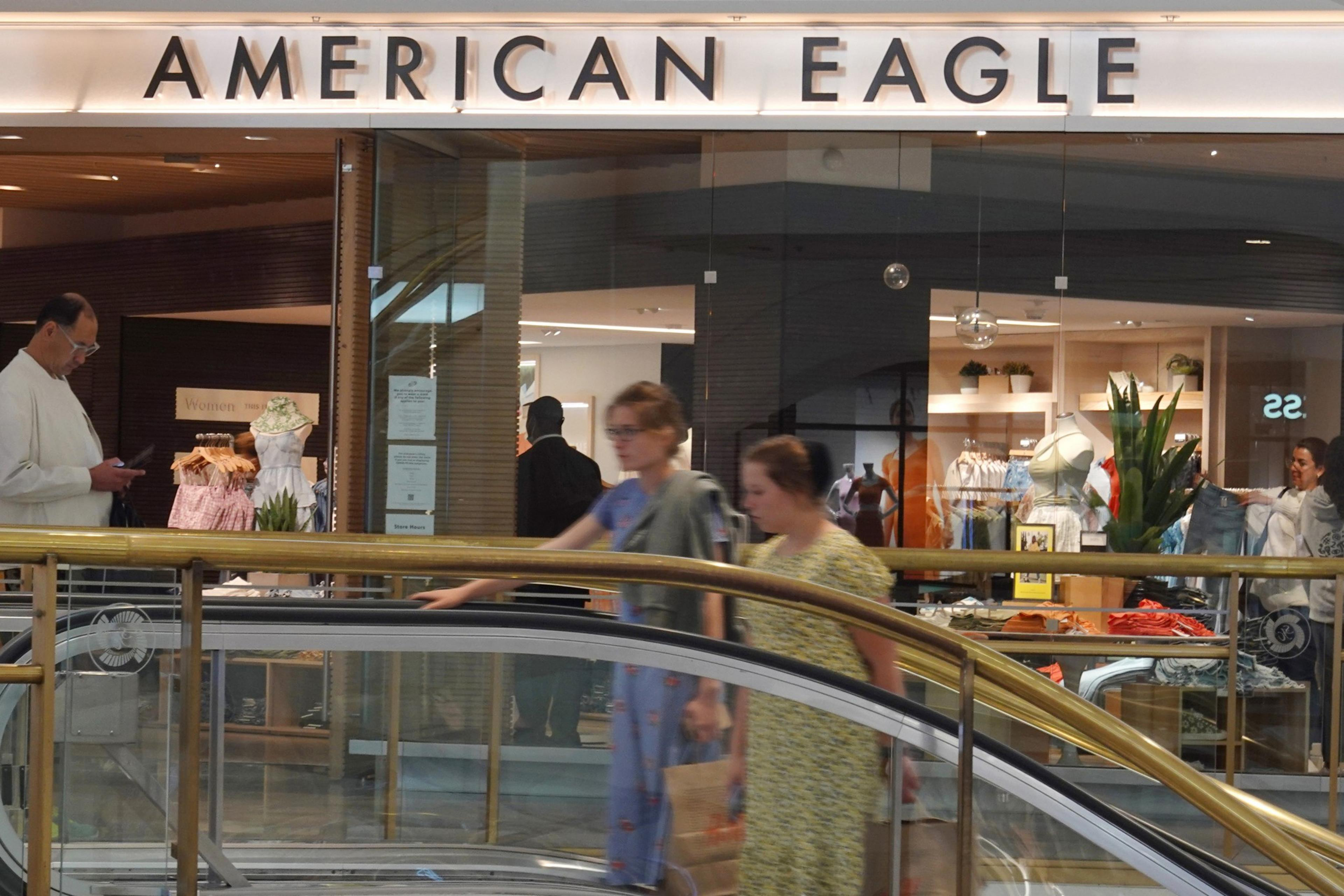 People are walking past an American Eagle store inside a mall, with a man reading on his phone.
