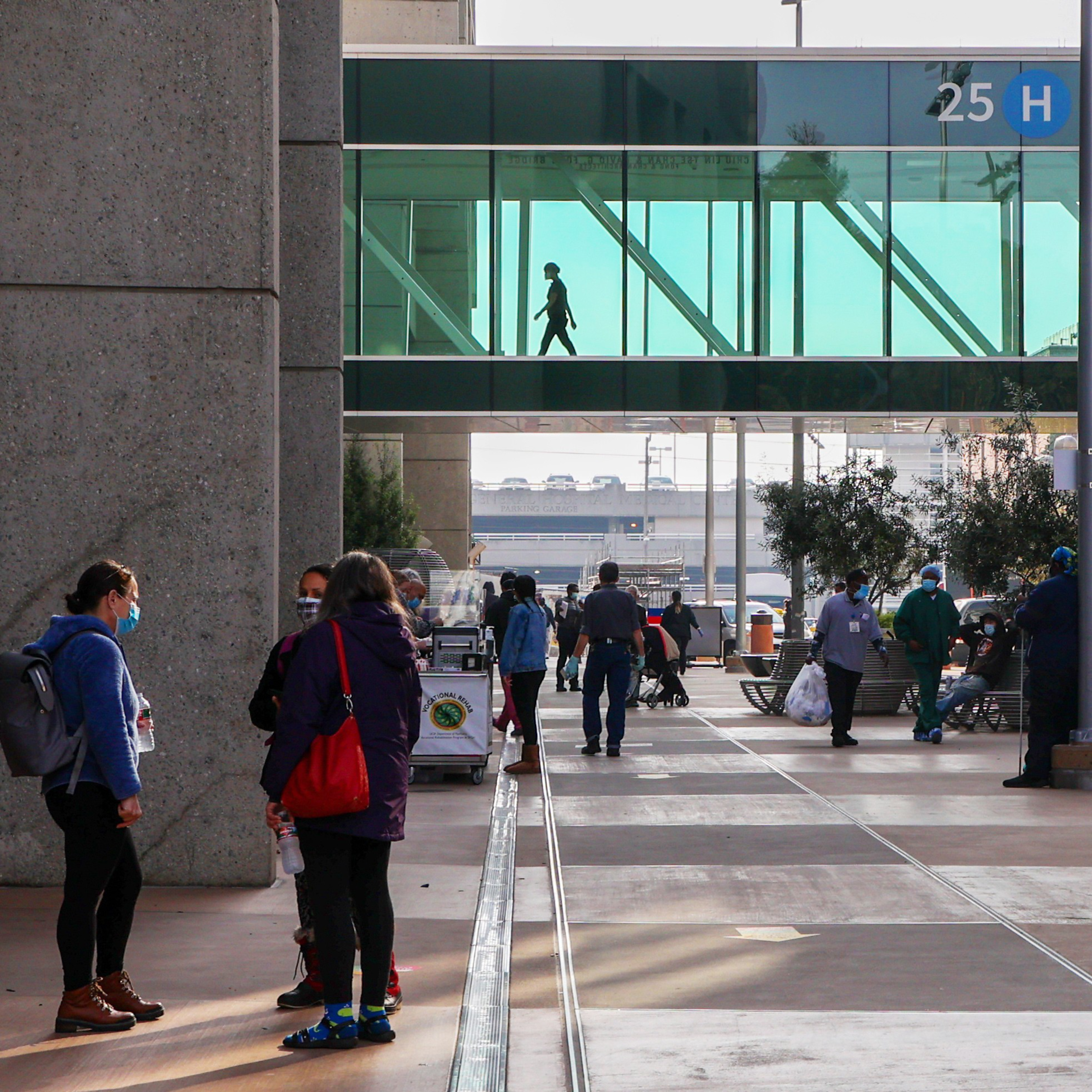 A person crosses a glass-walled bridge above a bustling outdoor terminal with passengers and a vendor cart.