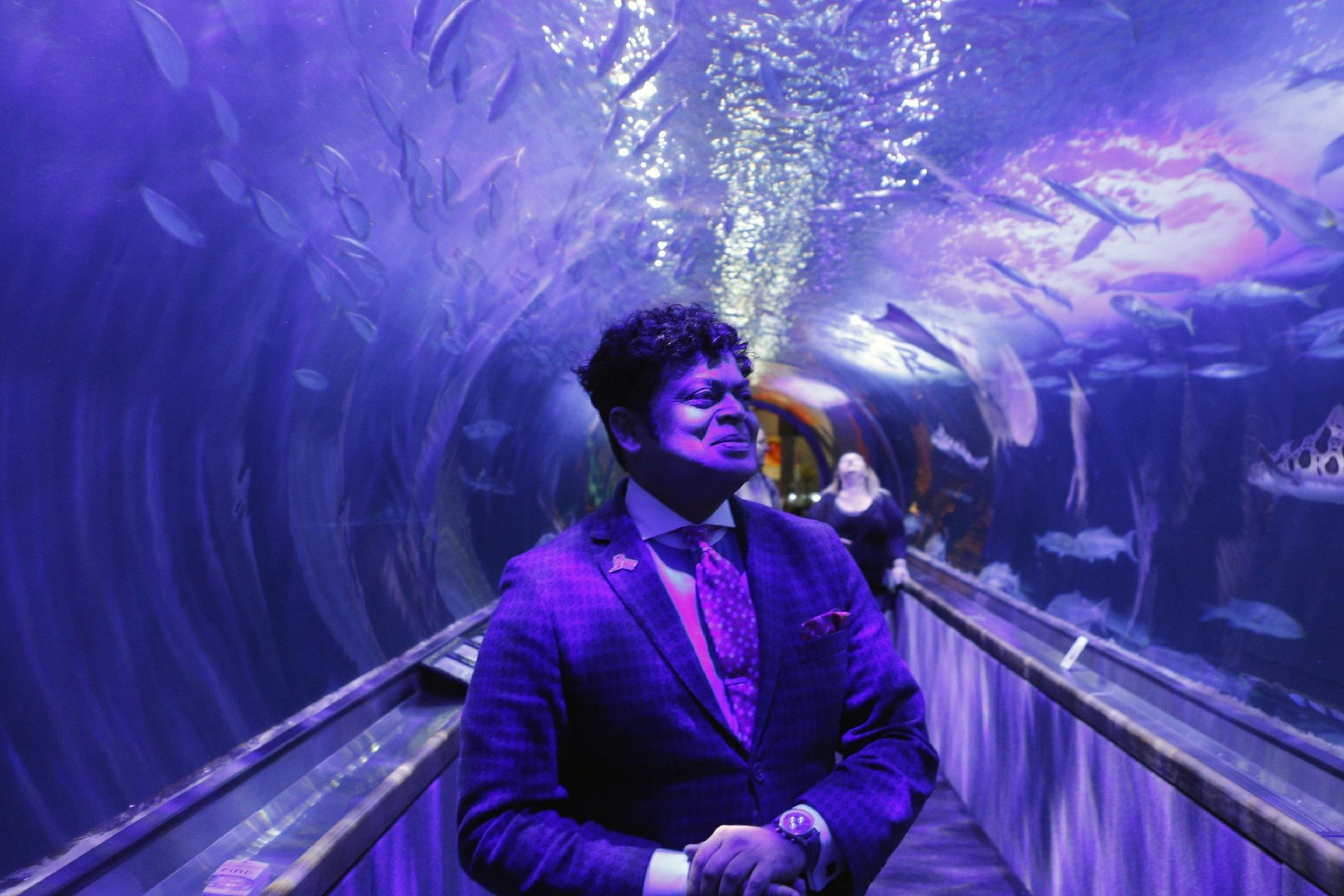 A man in a suit stands in an illuminated aquarium tunnel surrounded by swimming fish and a shark. The scene is bathed in purple and blue lighting.