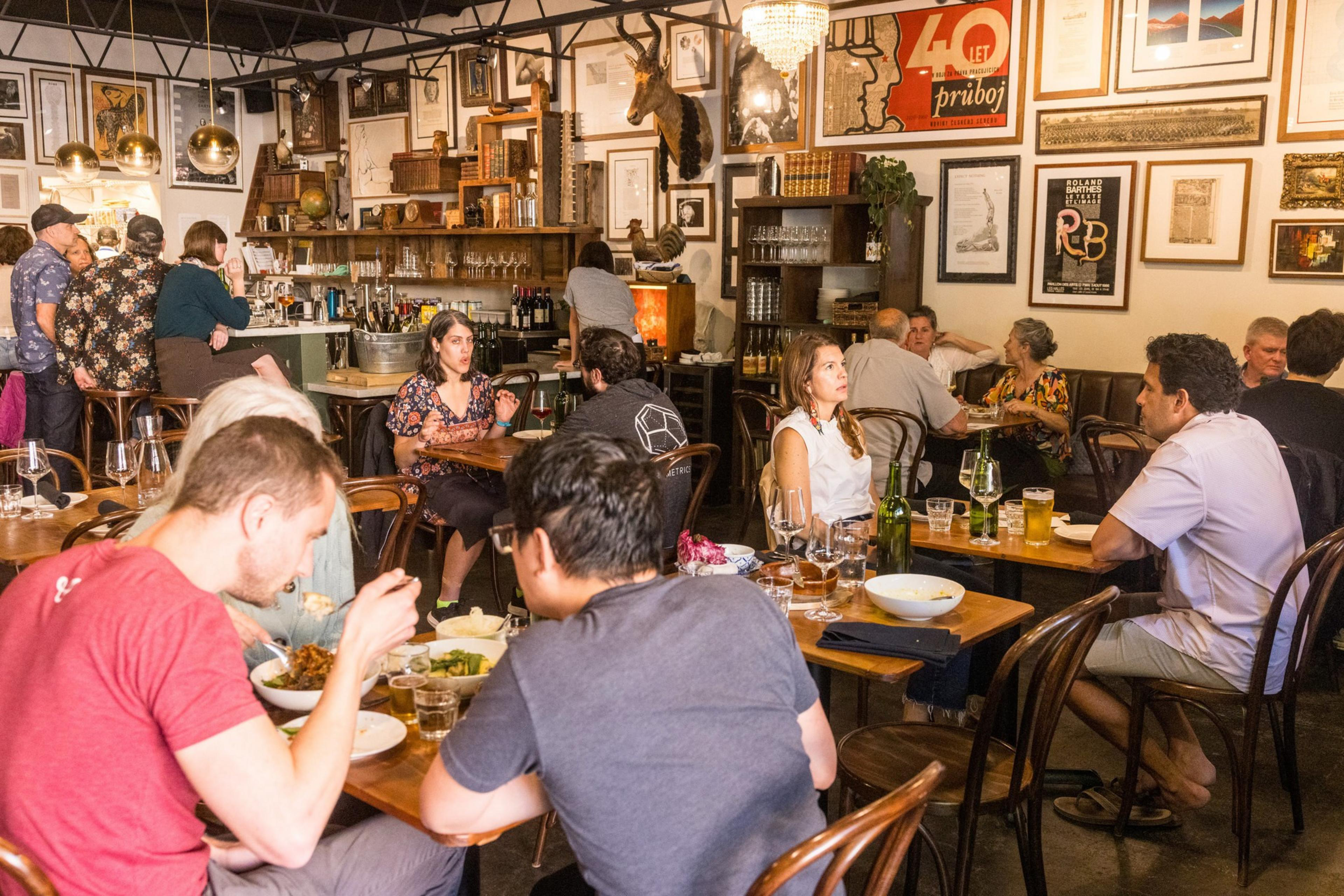 The image depicts a busy, warmly lit restaurant filled with patrons enjoying meals. People are seated at wooden tables, with artwork and decorations on the walls.