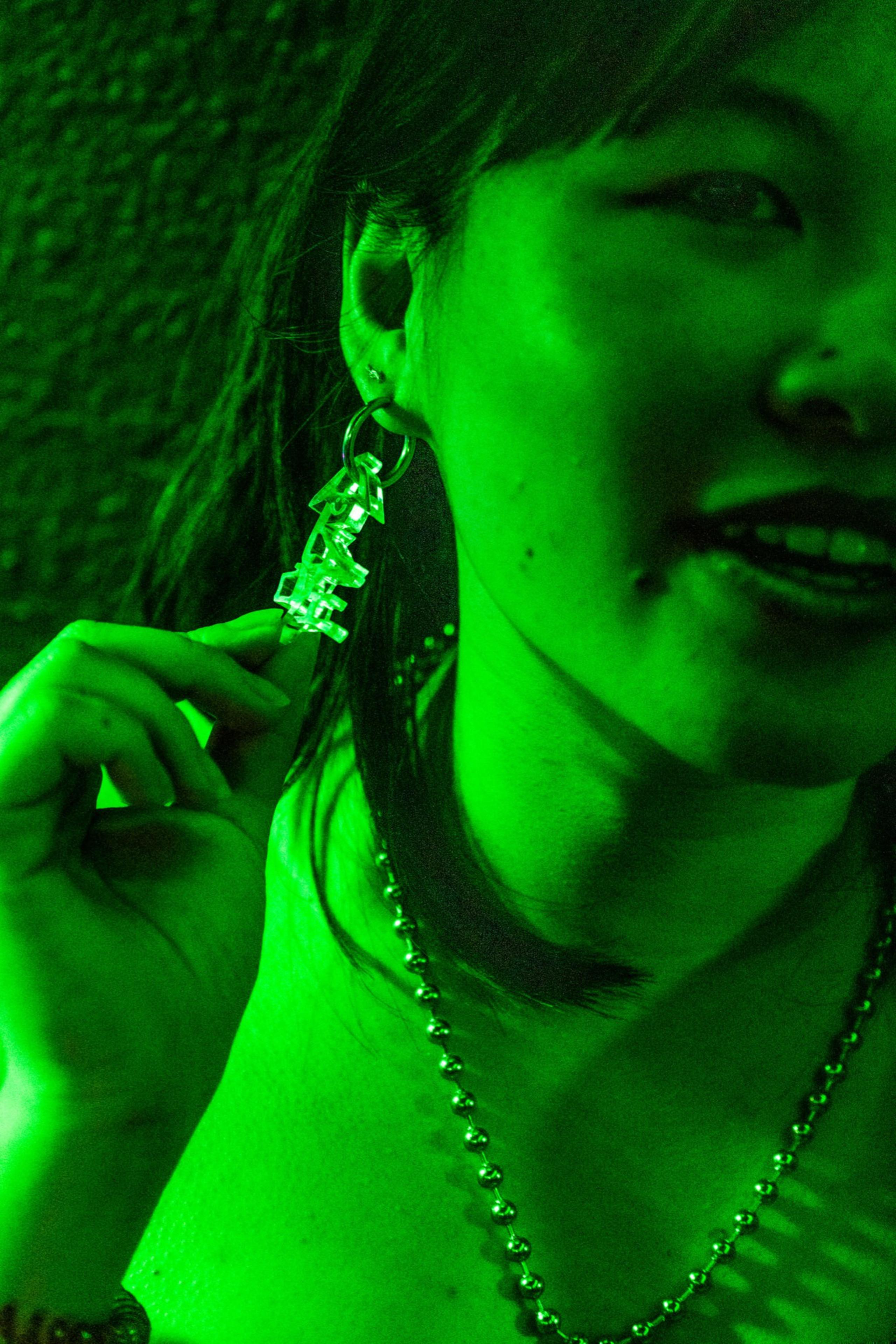 The image shows a person under green lighting, wearing large earrings and a necklace, with one hand touching the earring.