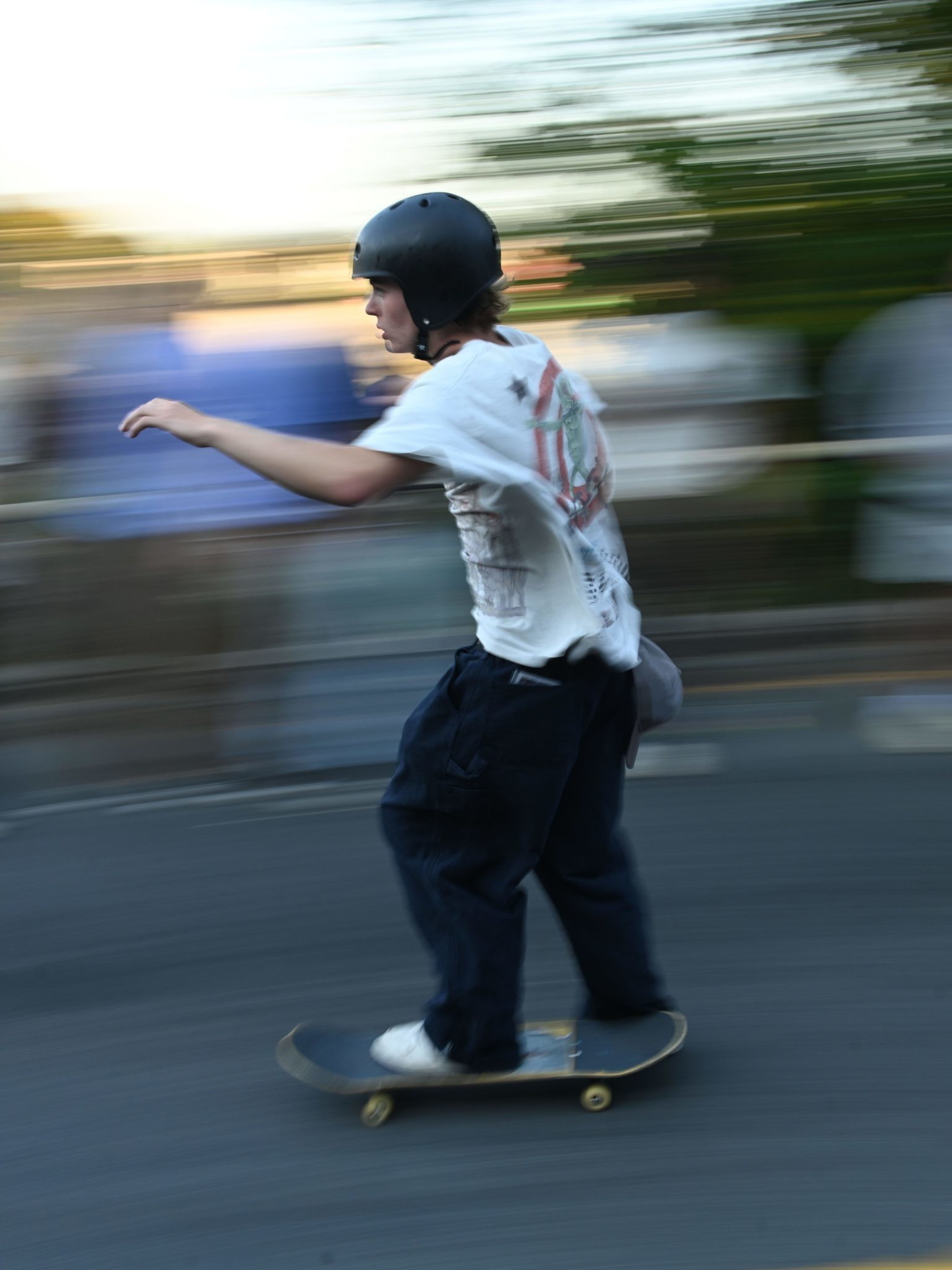 A person wearing a helmet rides a skateboard on a street, moving quickly past a blurred background of people and trees. The motion creates a dynamic, energetic feel.