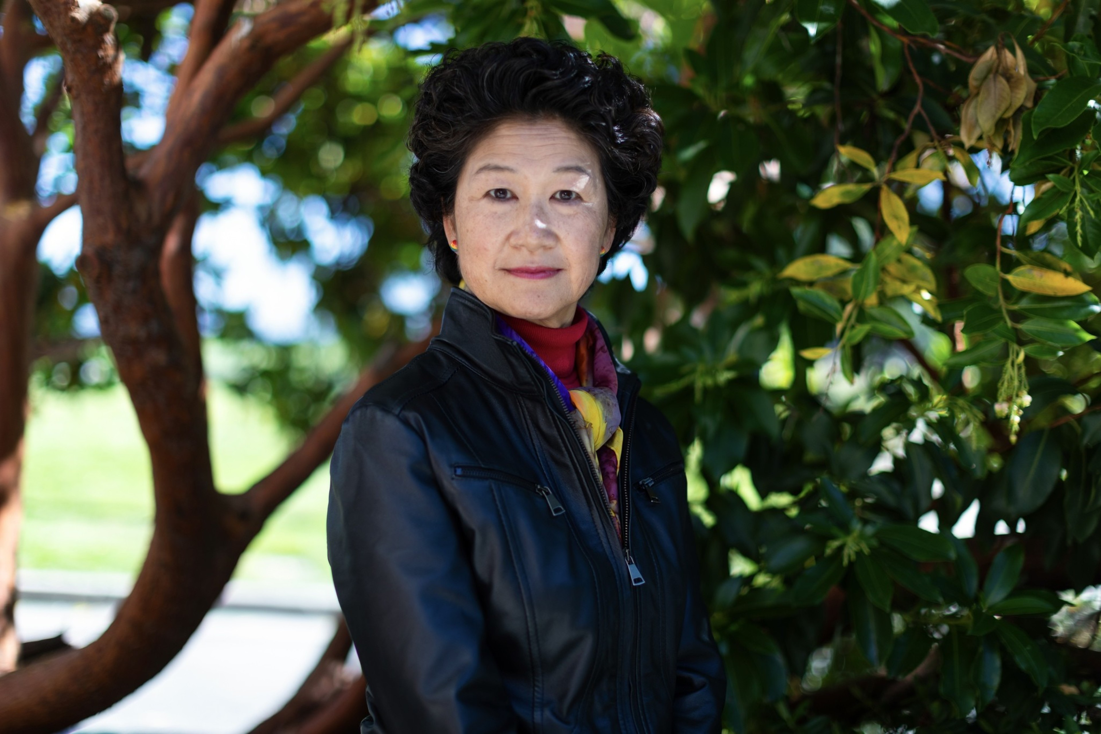 A woman in a black jacket stands outdoors near a tree with lush green leaves, looking at the camera with a neutral expression. There is sunlight filtering through the foliage.