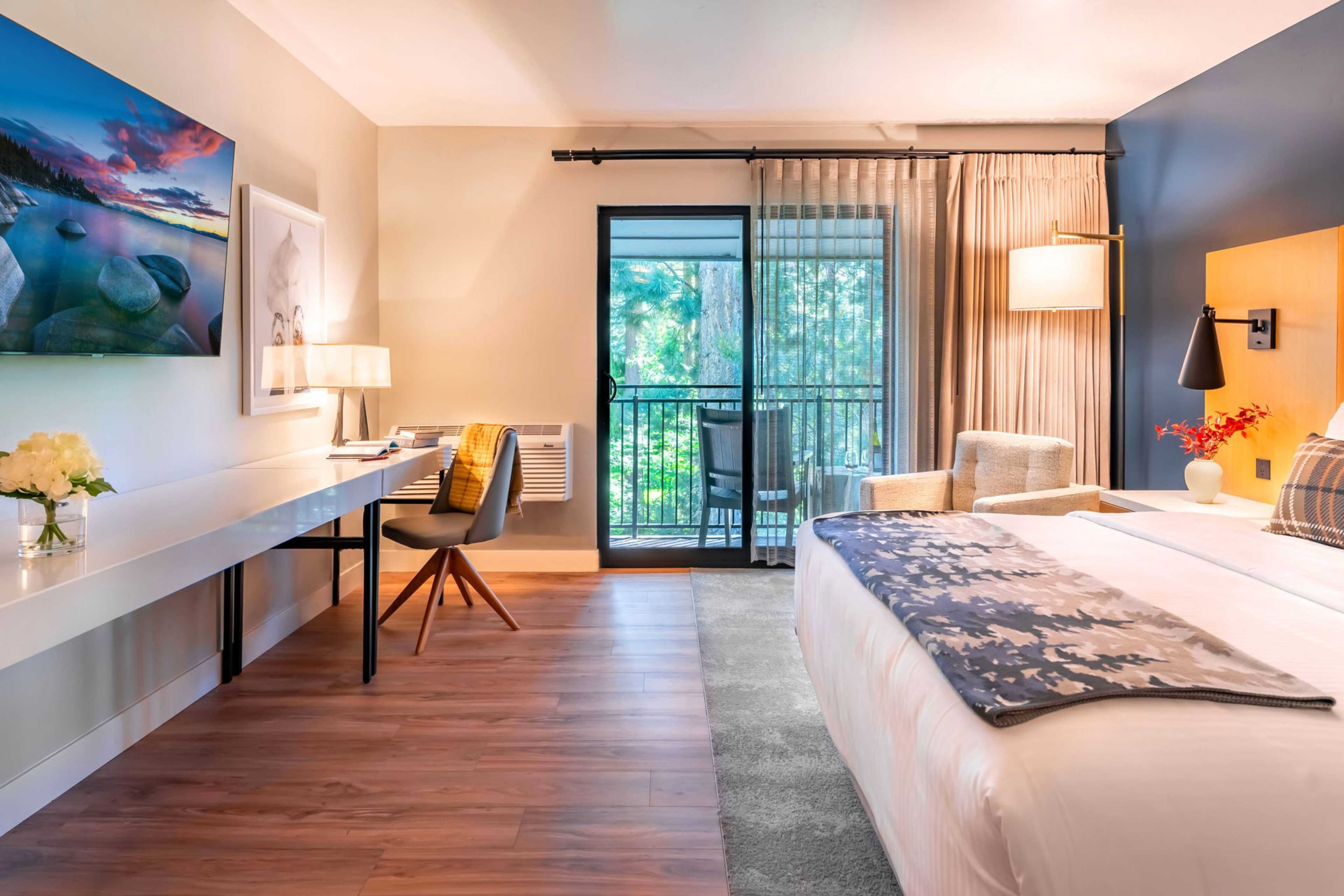 A room with a bed opposite a long table looks out to a balcony with trees in view