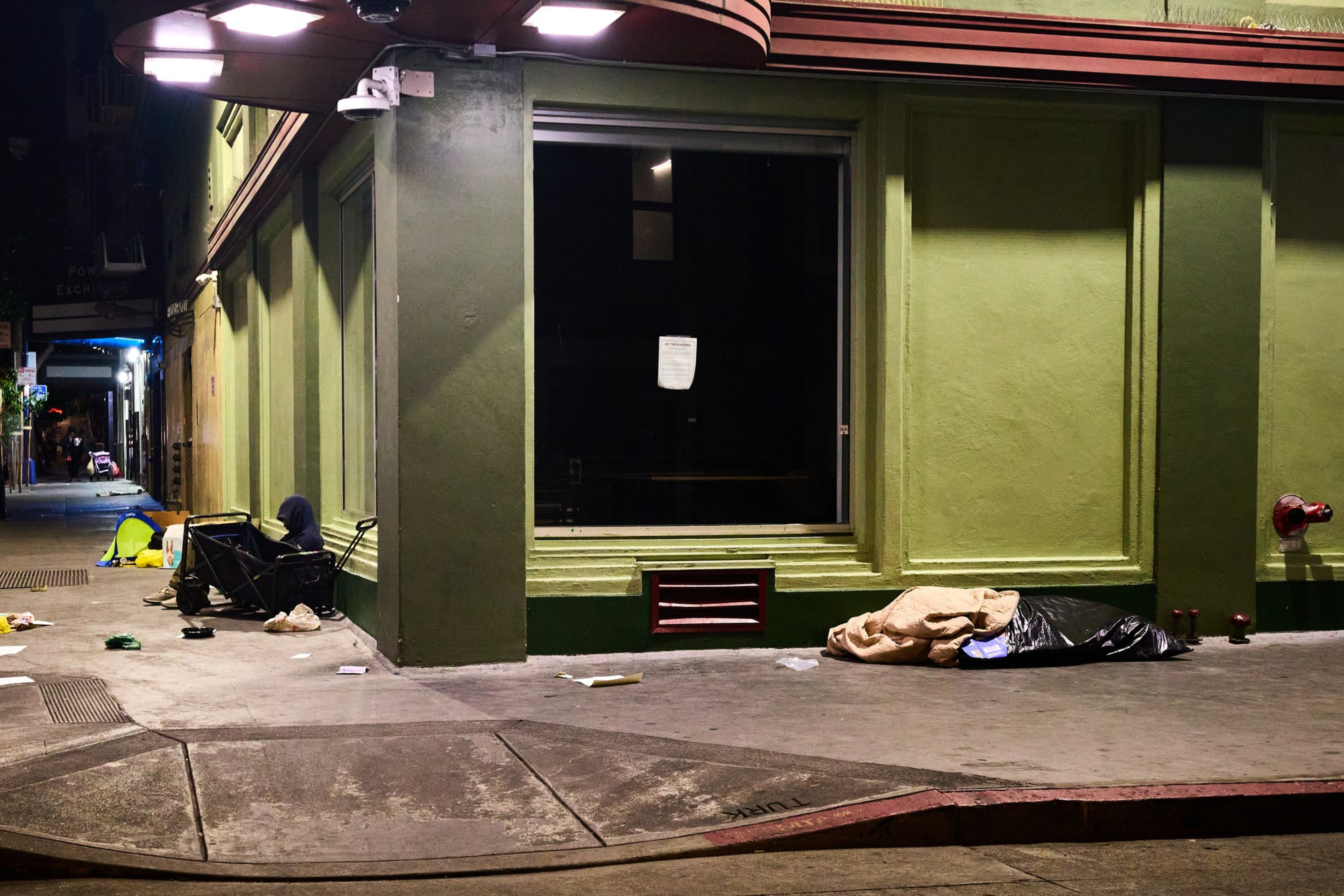 A dimly lit urban street corner at night shows a closed store with a large window, a person sitting against the wall, and scattered belongings on the sidewalk.