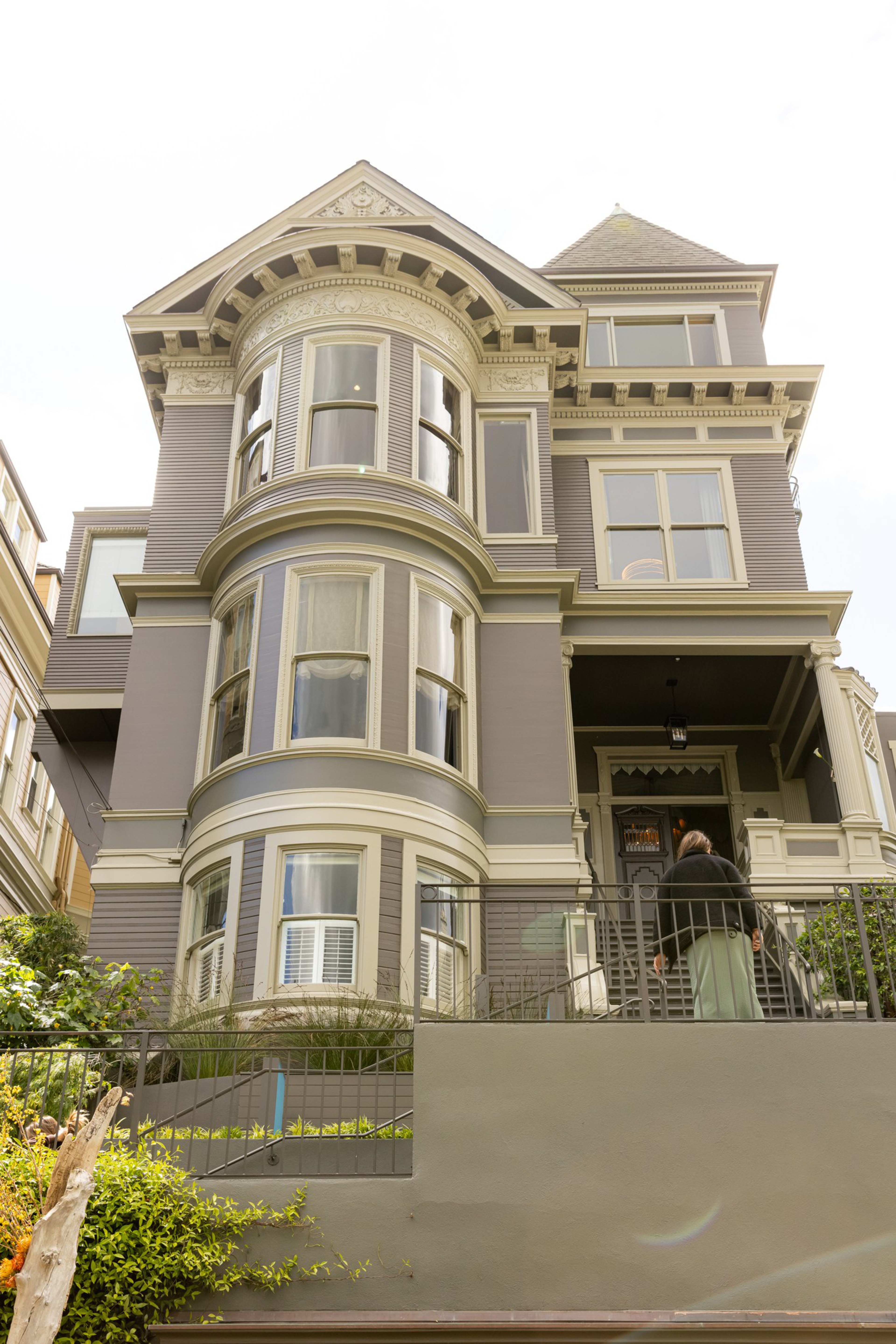 A large, ornate Victorian-style house painted gray with white trim features three rounded bay windows and a porch with a person walking up the stairs.