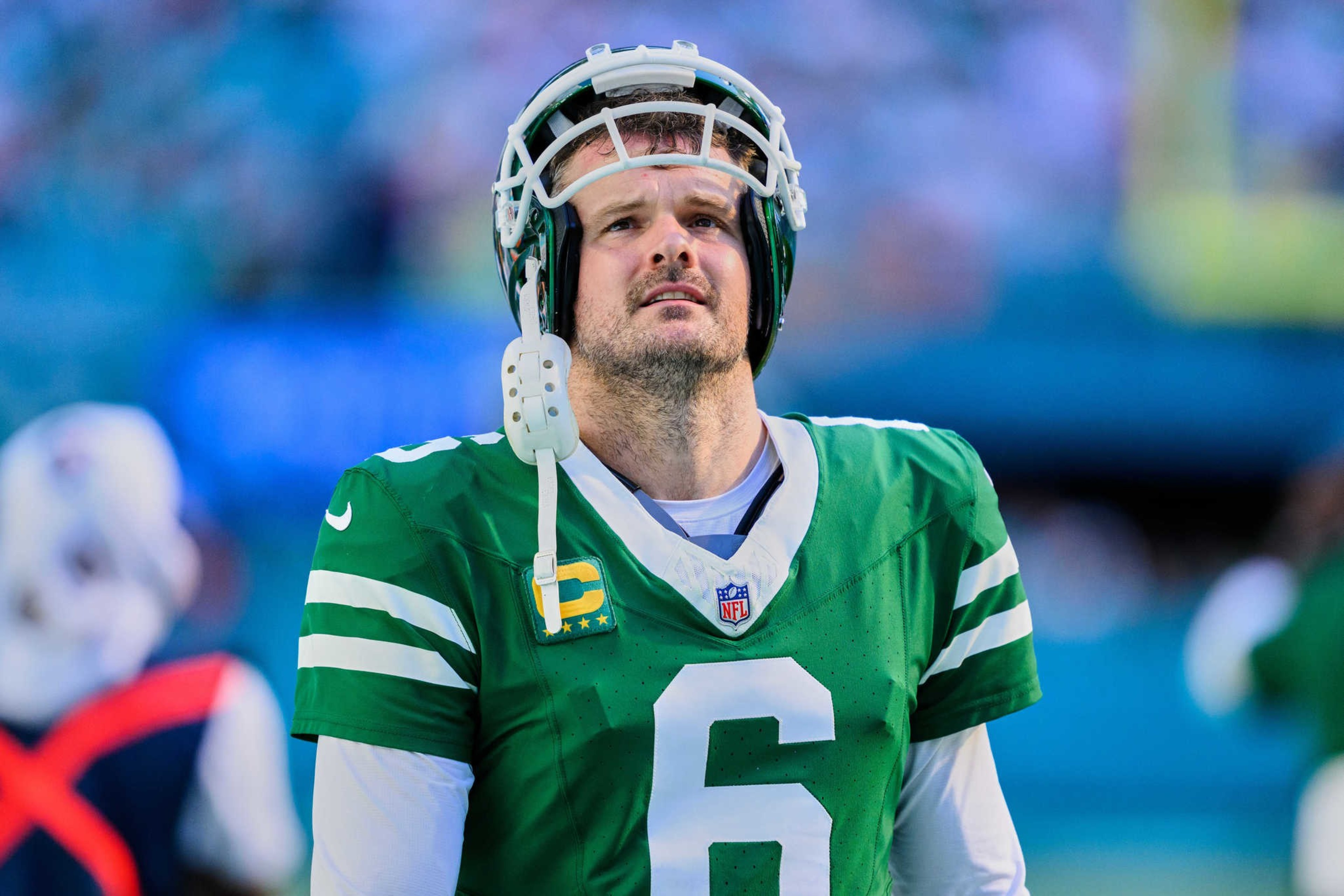 A football player in a green jersey with the number 6 looks upward. He wears a helmet, and the background is a blurred stadium.
