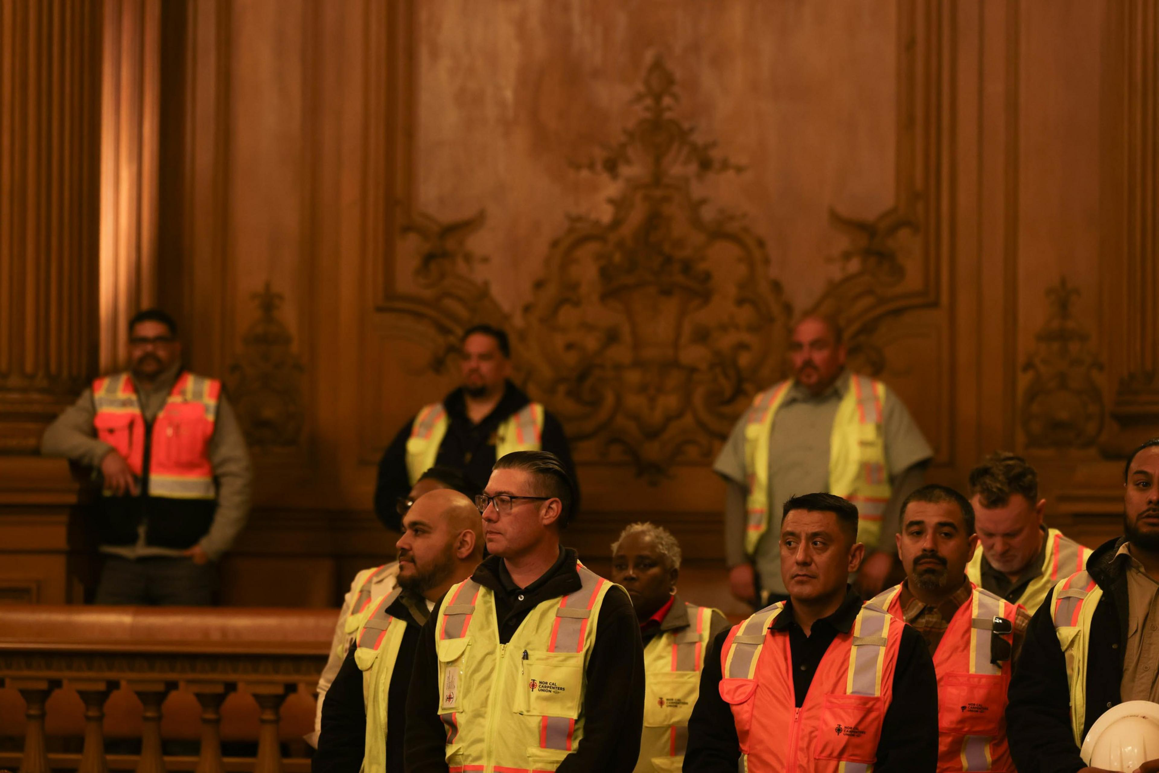 A group of people wearing reflective vests stands indoors against an ornately carved wooden wall. They appear attentive and focused.