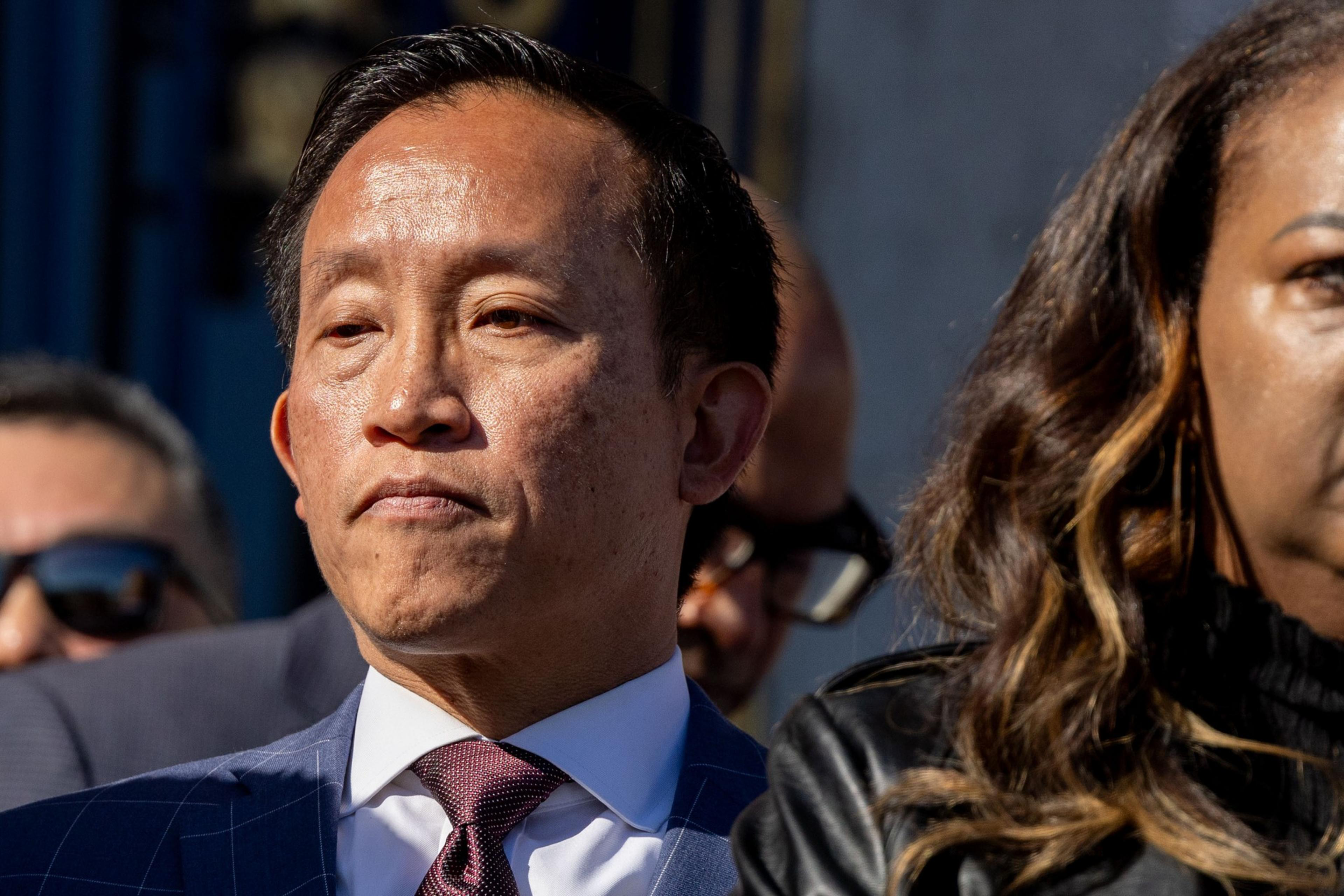 A man in a suit with a serious expression stands outdoors, partially lit by sunlight. Another person, partially visible, stands beside him.