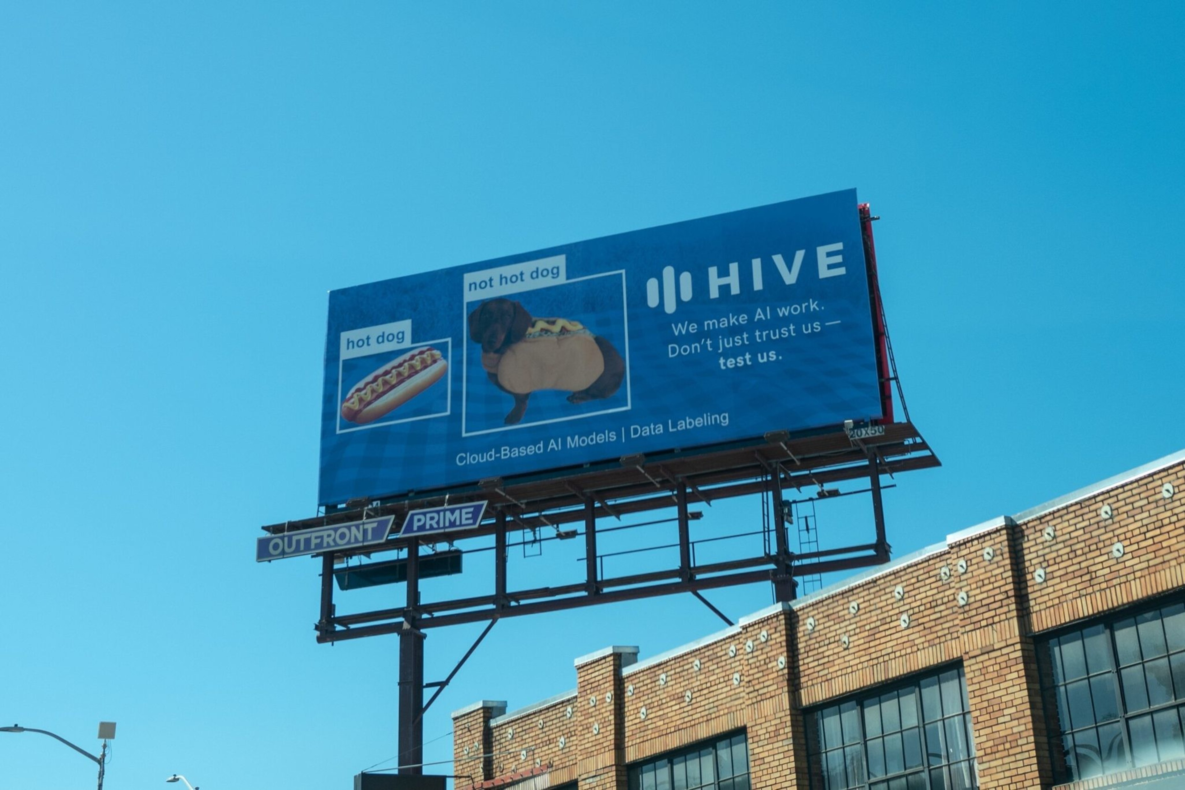 A billboard displays images labeled &quot;hot dog&quot; (actual hot dog) and &quot;not hot dog&quot; (dog in hot dog costume) promoting Hive's AI services with the tagline &quot;We make AI work.&quot;