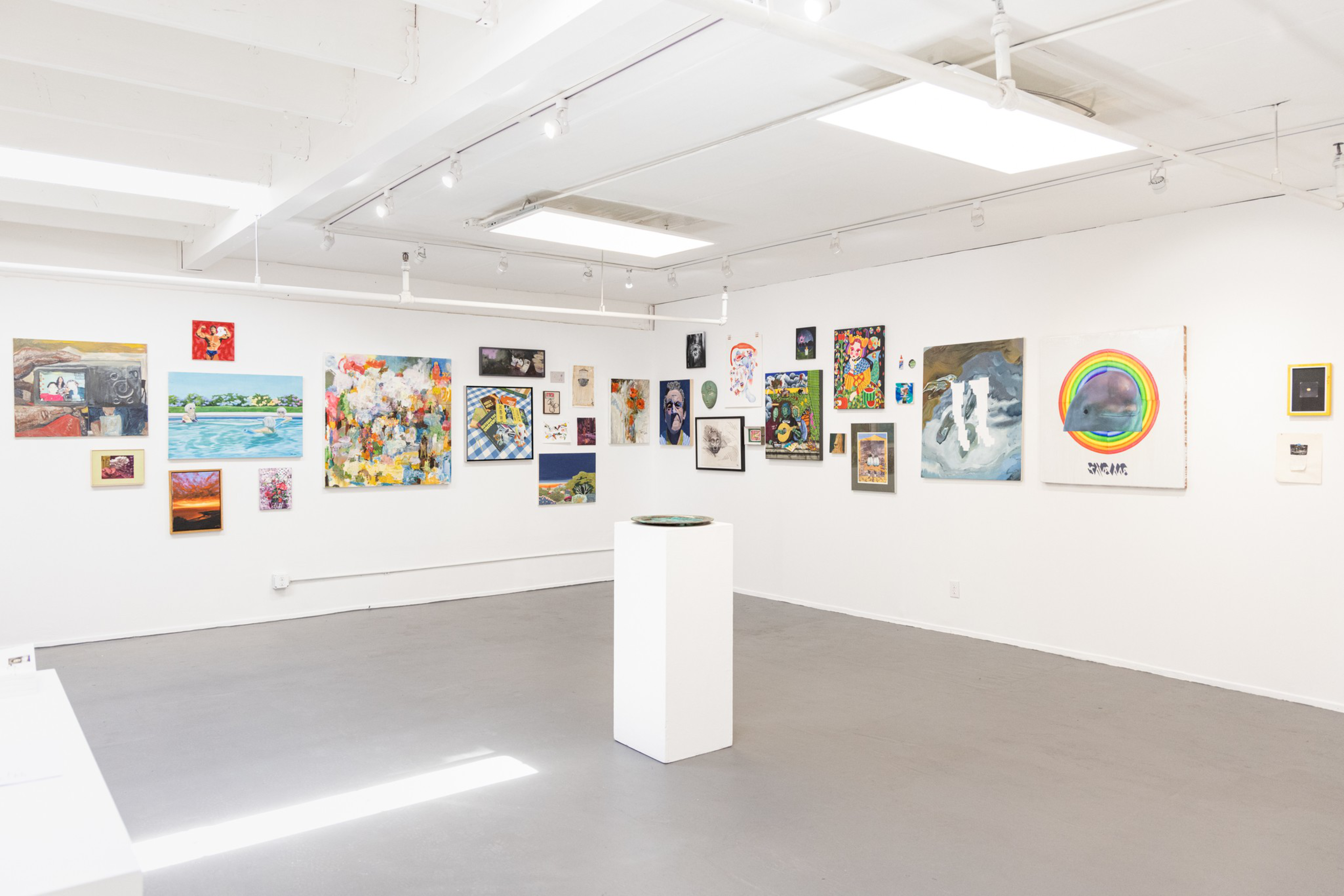 A bright gallery room displays various colorful and monochrome artworks on white walls surrounding a white pedestal in the center of the gray floor.