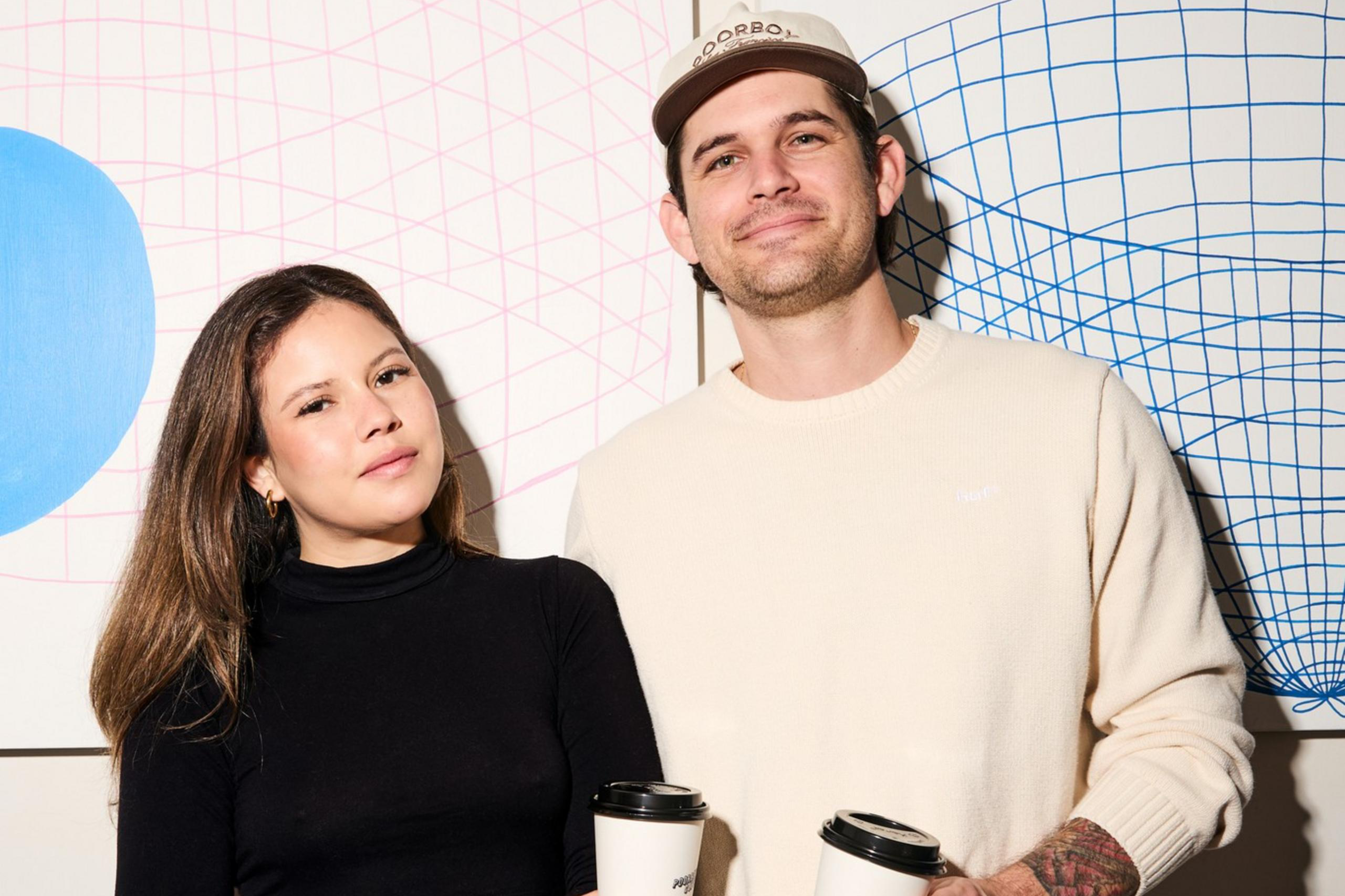 A man and woman stand smiling, holding coffee cups. The woman wears a black top and jeans, while the man wears a cream sweater and cap. Geometric art is behind them.