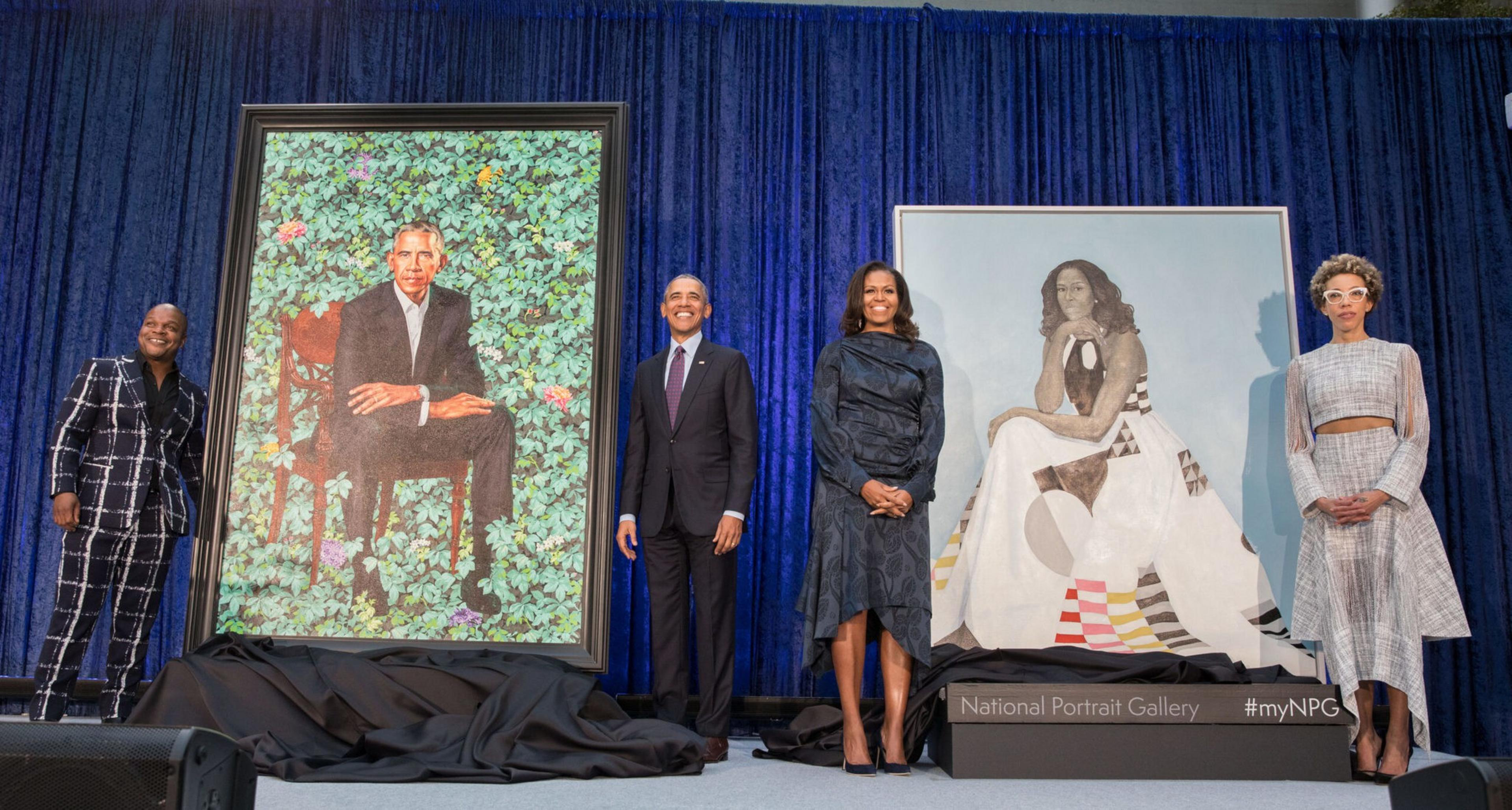 Four people stand on a stage between two large painted portraits of Barack and Michelle Obama, with a blue curtain backdrop.