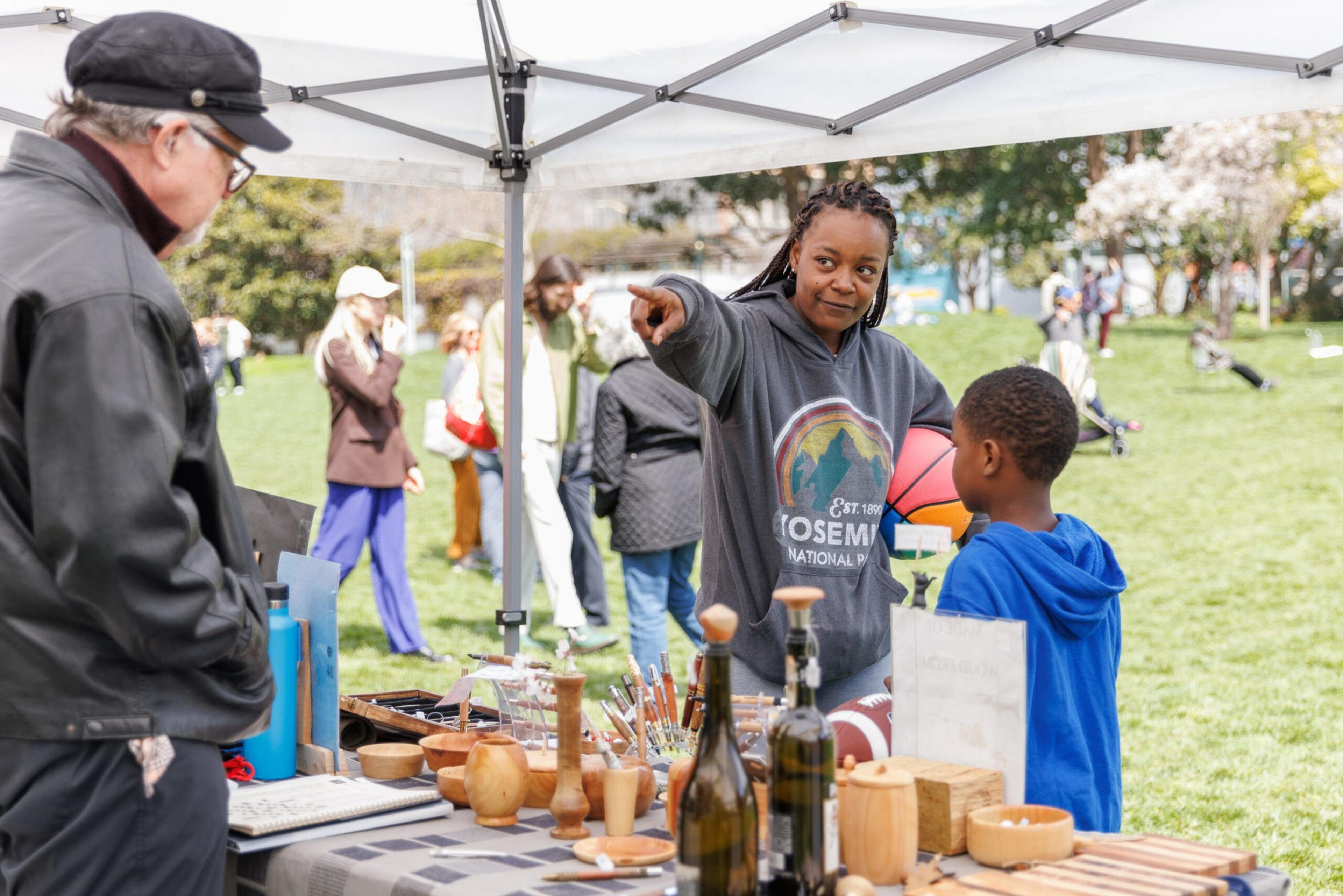 A woman at a market stall is interacting with a young boy, while people browse and chat in a grassy area with trees.
