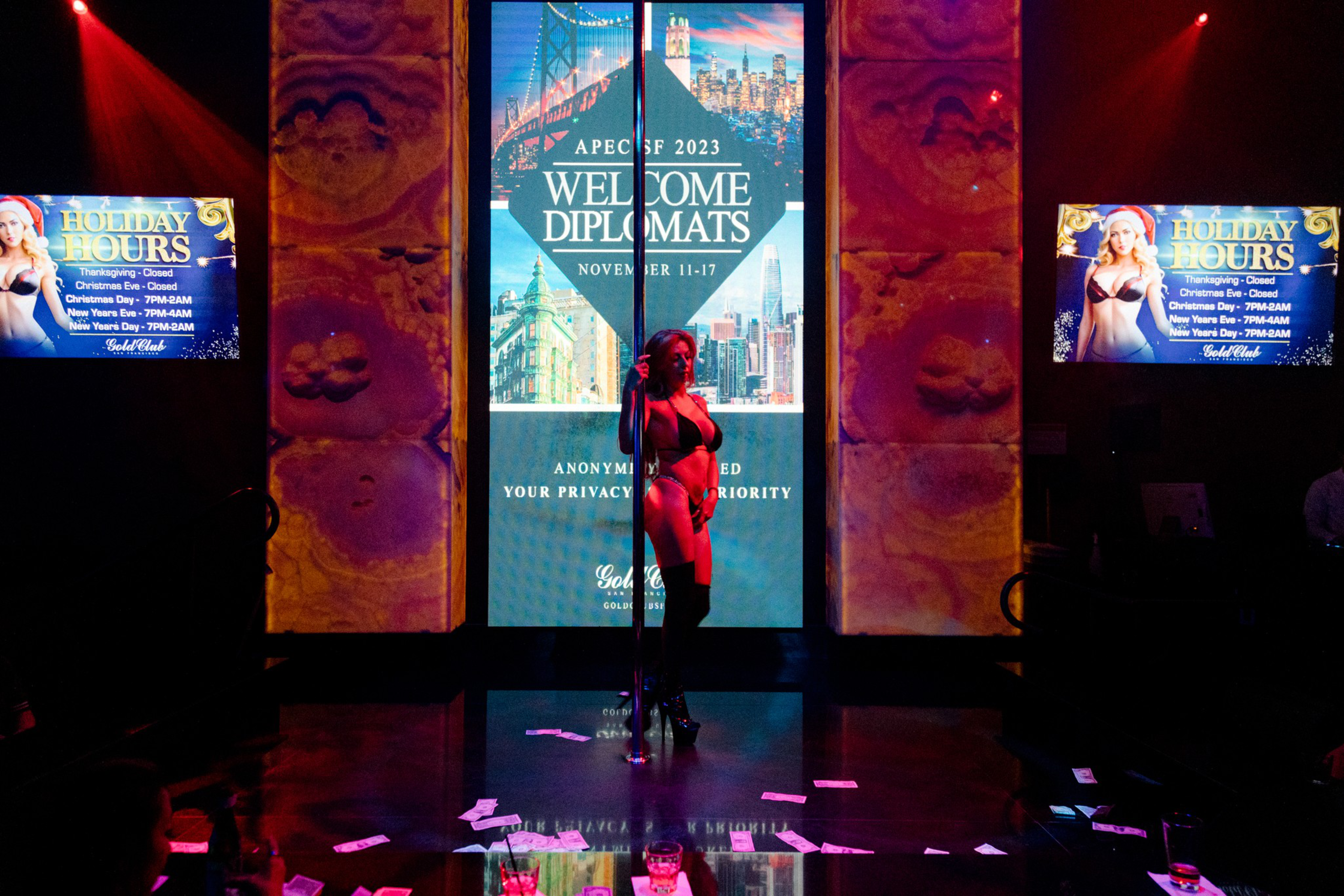 An exotic dancers stands in the middle of the dance floor with dollar bills on the ground and a LCD screen that reads "Welcome Diplomats - APEC SF 2023 - November 11-17"
