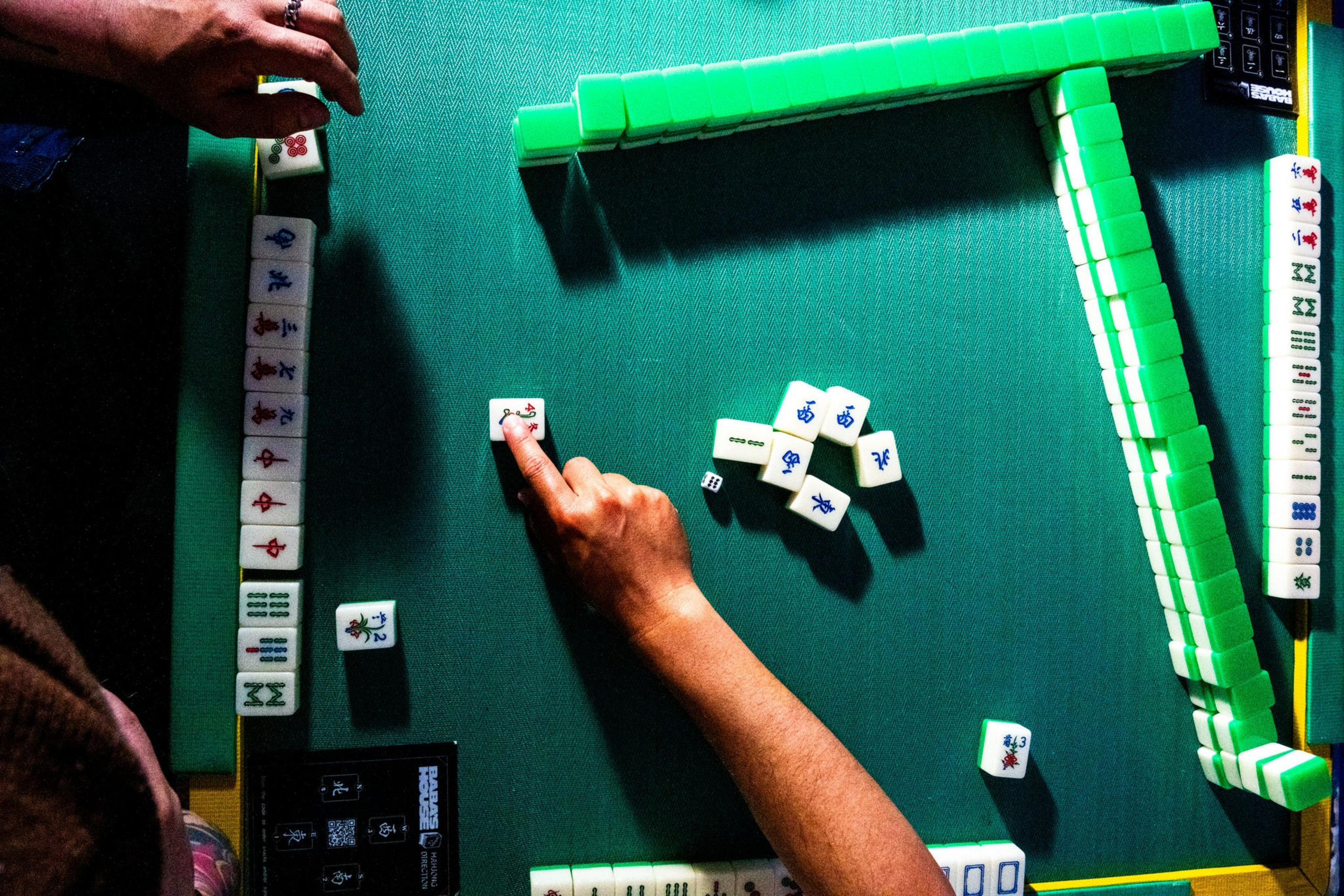 A Mahjong game is in progress on a green table. A hand points to a tile, while other tiles are arranged in lines. The game appears to be partway through.